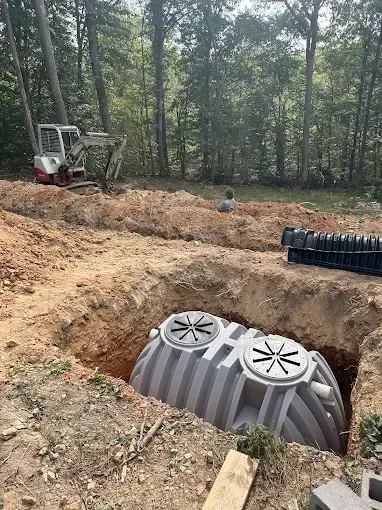 Septic tank being installed in a dirt trench with an excavator and worker in a wooded area.