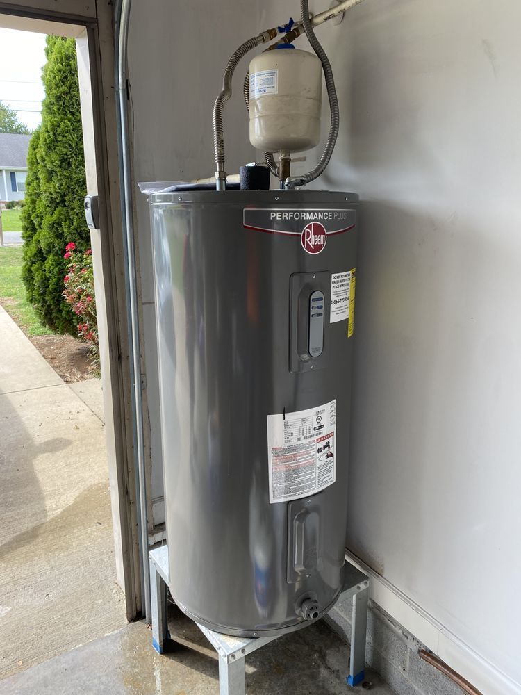 A Rheem water heater in a garage, with an expansion tank on top, sitting on a metal stand.