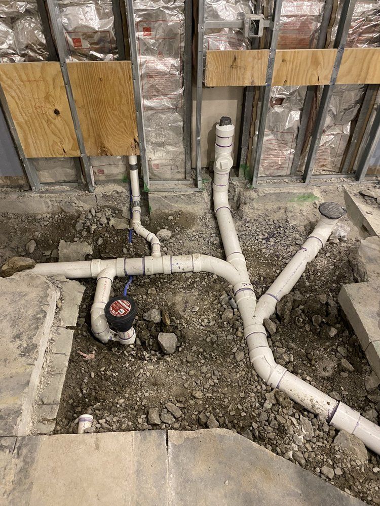 Plumbing pipes in a construction site, with open wall, unfinished concrete, and gravel on the ground.