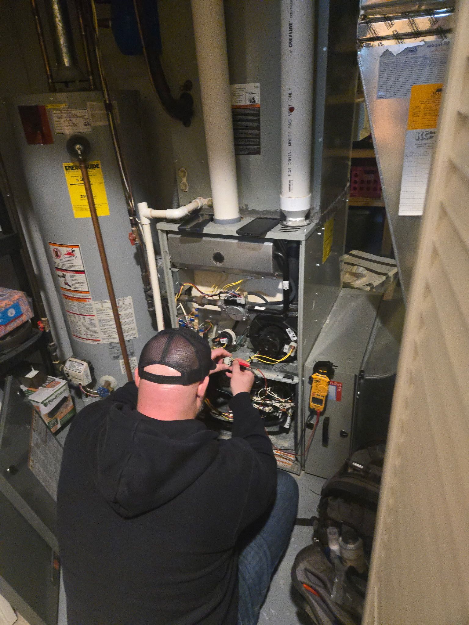 Technician inspecting electrical panel in a basement utility room, with exposed wiring and pipes.