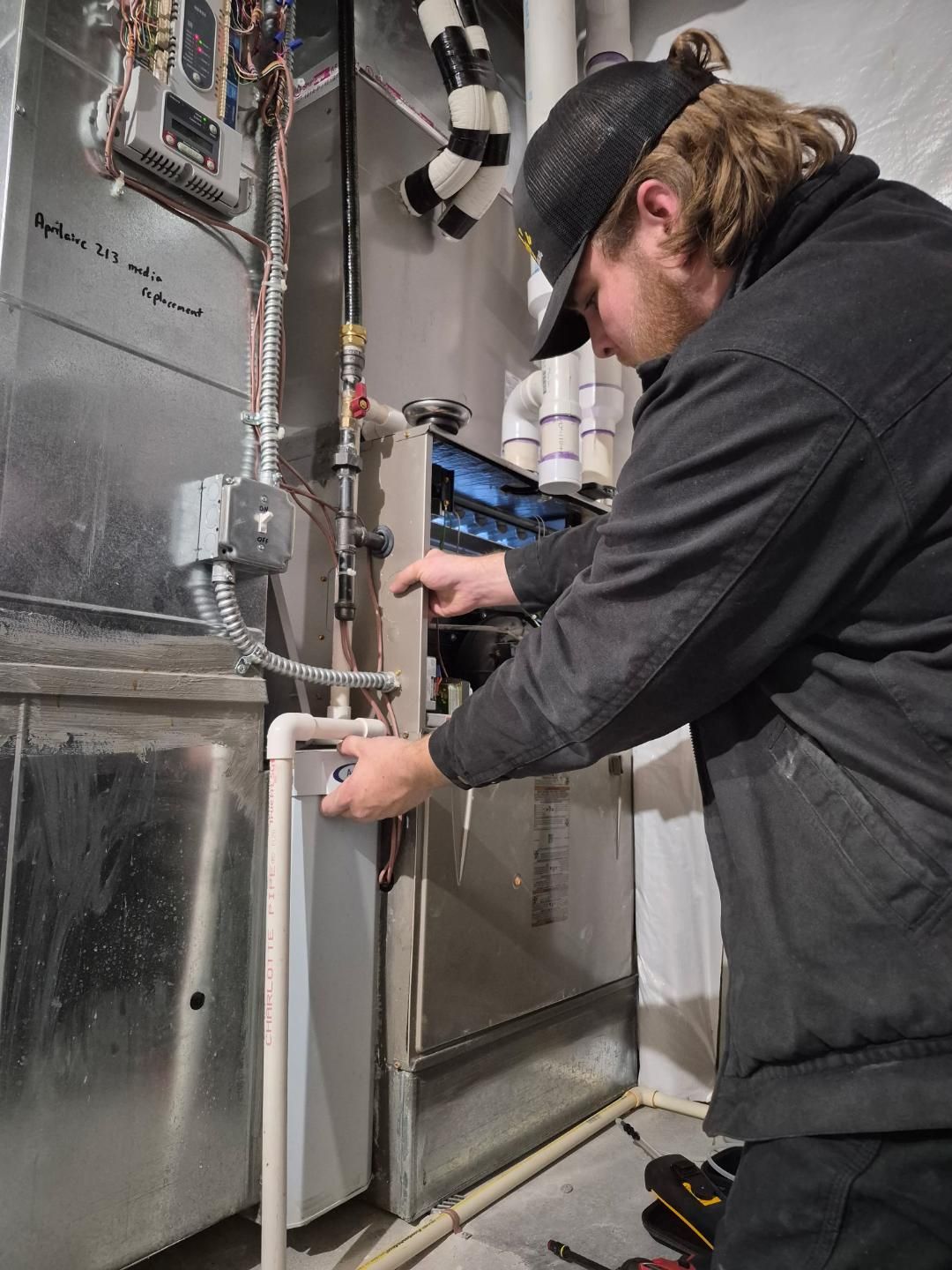 Technician inspecting electrical panel in a basement utility room, with exposed wiring and pipes.