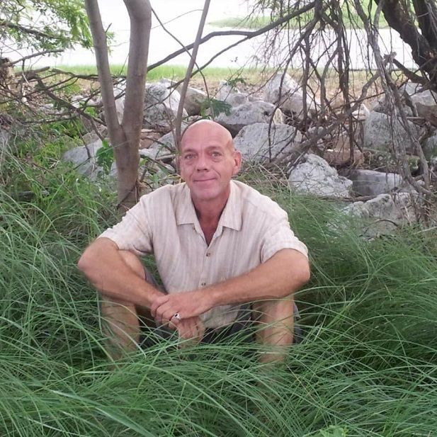 Man sitting in tall grass outdoors near trees and rocks, looking at the camera.