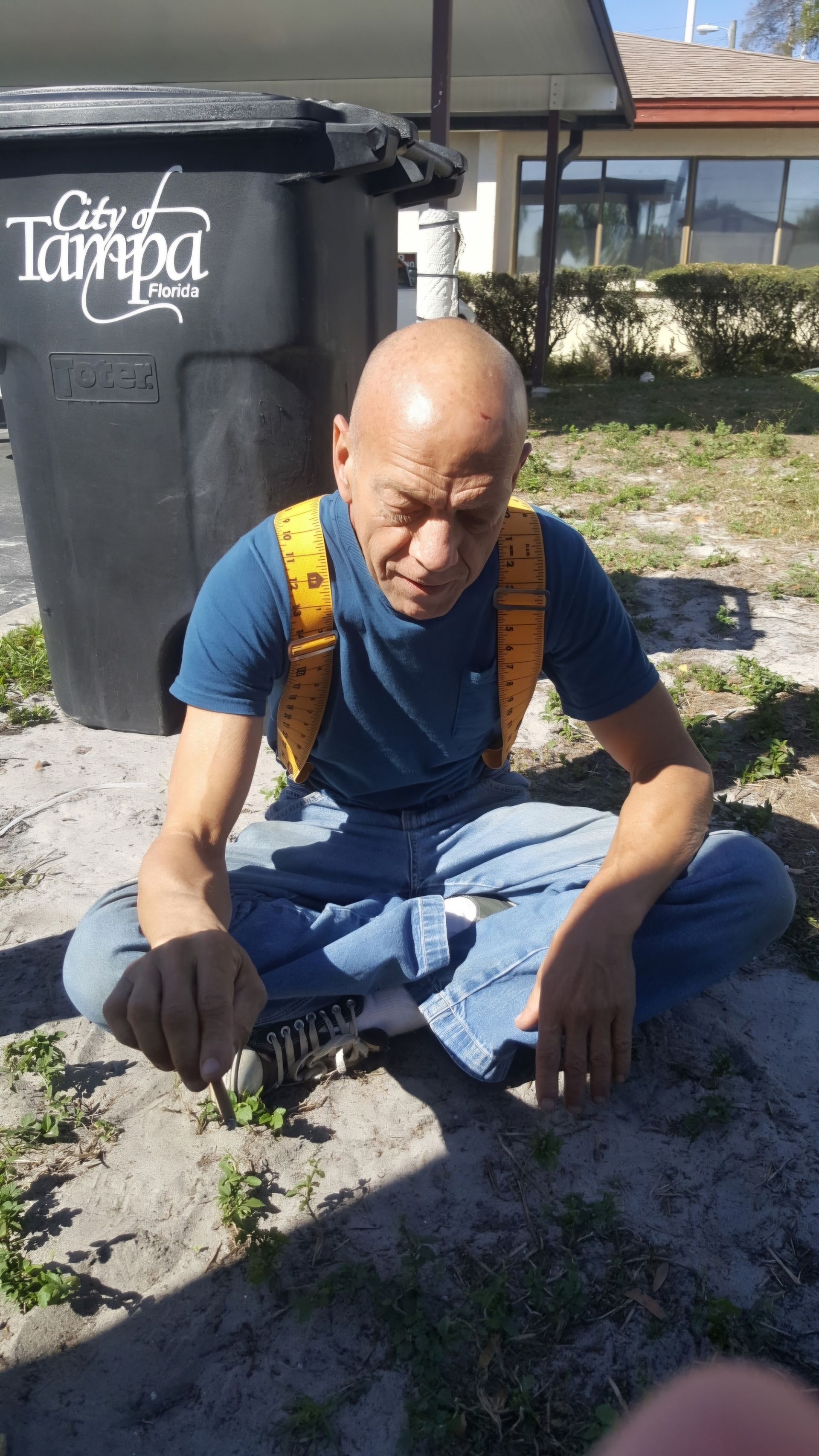 Person sitting on the ground beside a trash bin in sunlight, wearing a blue shirt and yellow backpack straps.