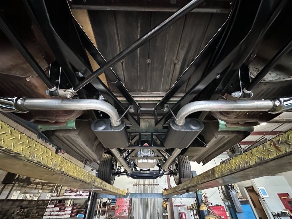 Underside view of a vehicle on a lift, showing exhaust system, chassis, and wooden bed.