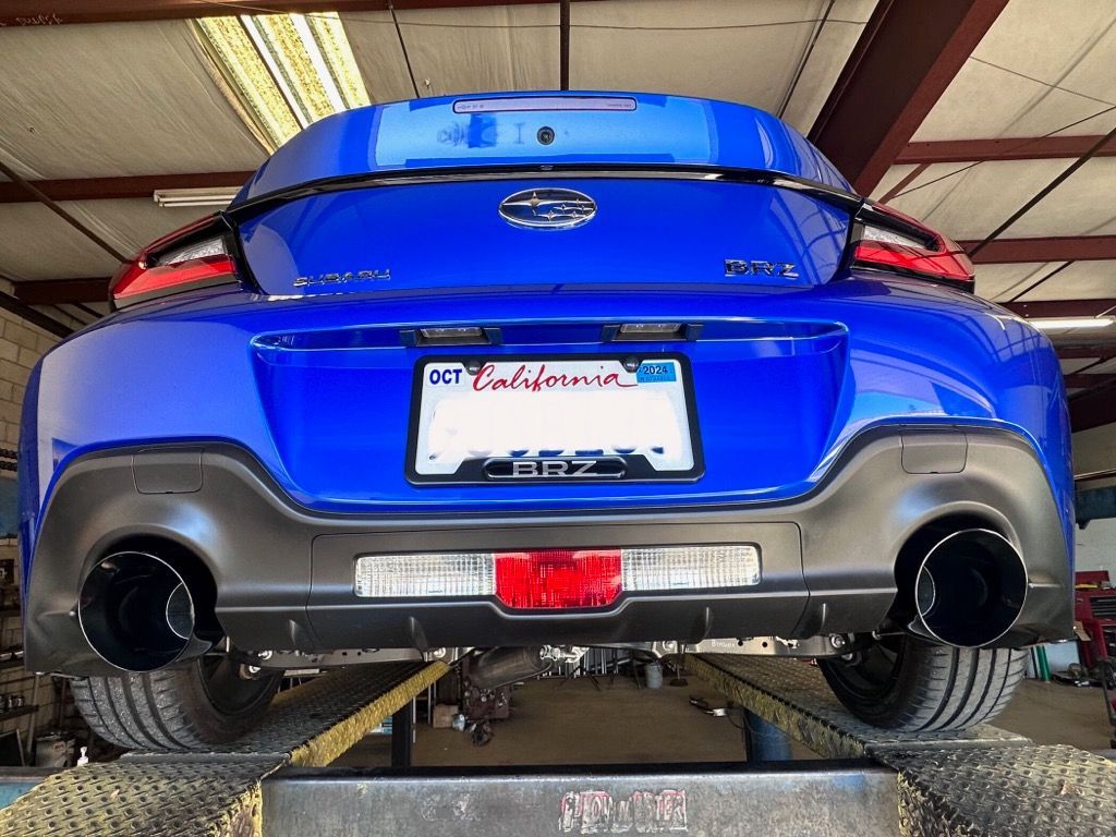 Blue sports car's rear, elevated on a lift, showcasing black exhaust pipes and California license plate.