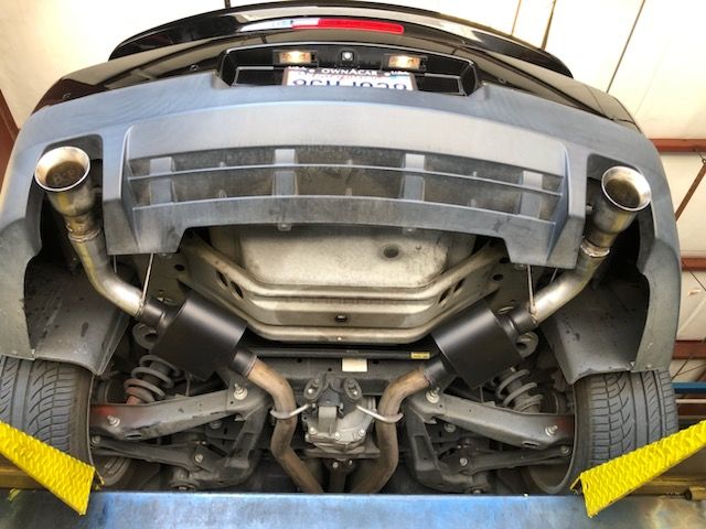 Underside of a black car on a lift showing an aftermarket dual exhaust system with black mufflers and polished tips.