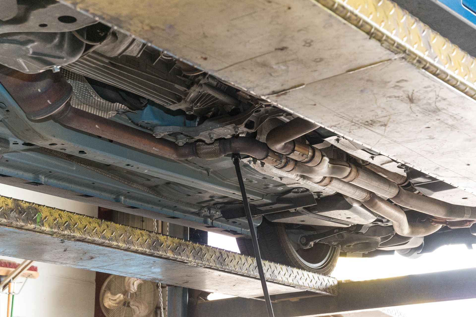 Underside of a car on a lift; oil draining from a dark pipe. Metal and exhaust components are visible.