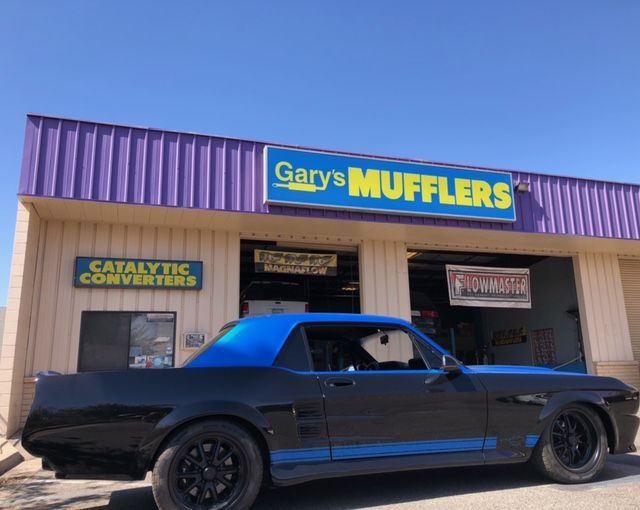 Black muscle car with blue stripes parked in front of Gary's Mufflers shop. Blue and purple building.