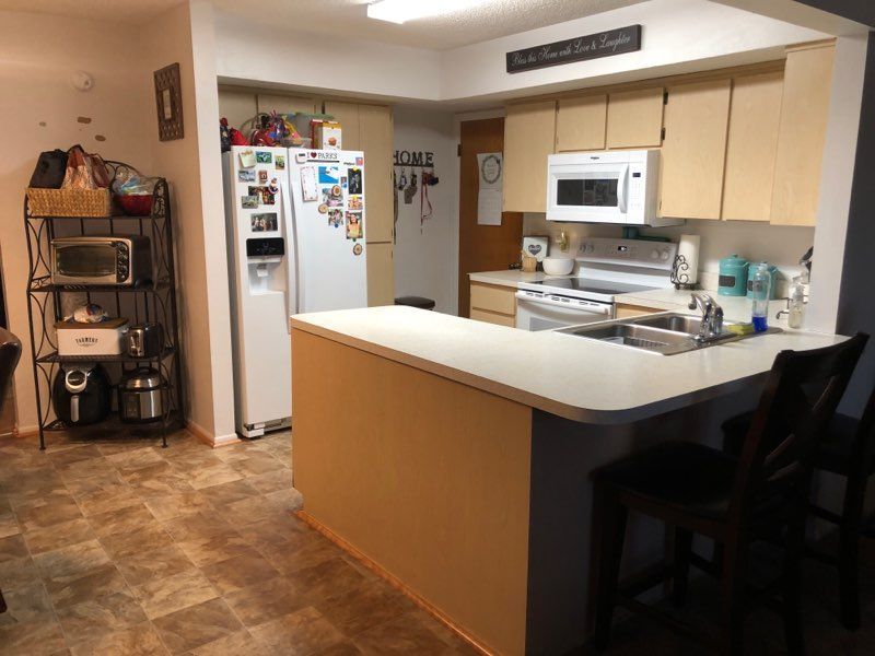 A kitchen interior with light wood cabinets, white appliances, a peninsula counter with stools, and a metal shelving unit.