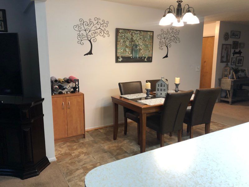 A dining area with a wooden table, dark chairs, a small cabinet, and wall-mounted tree art under a light fixture.