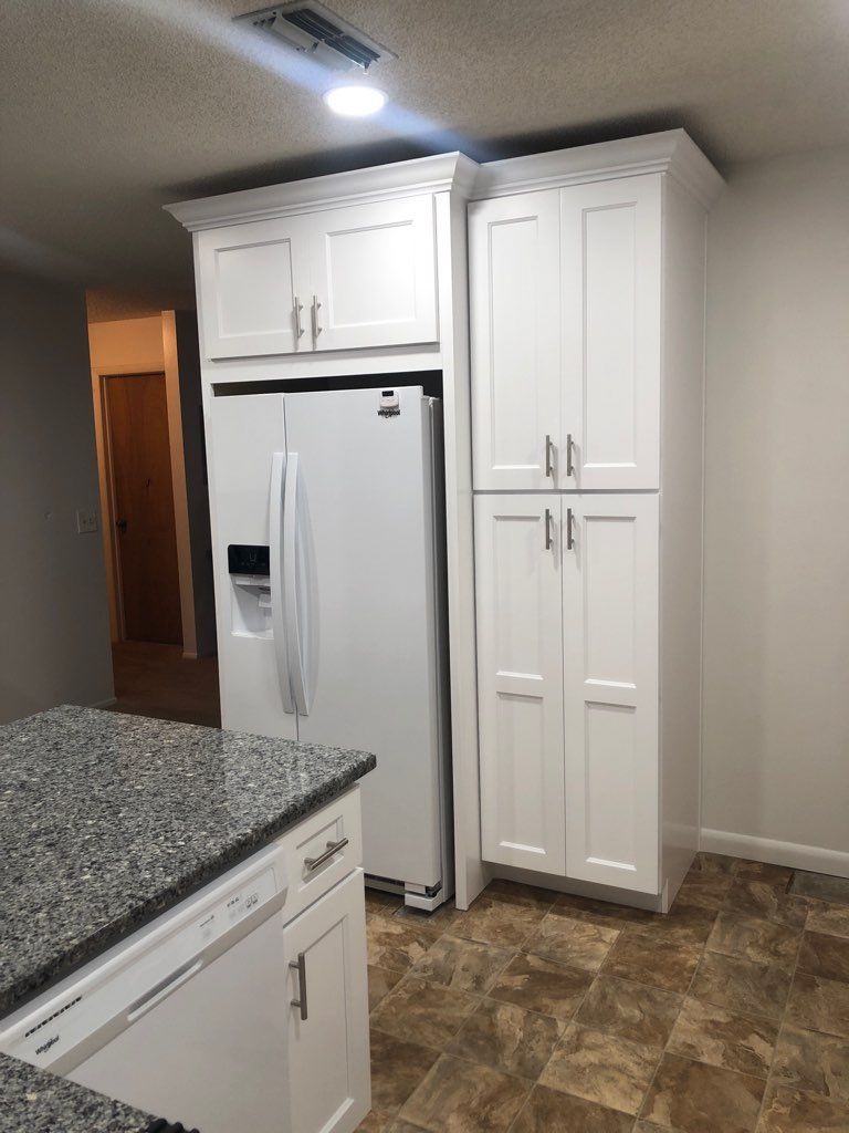 A white refrigerator beside a tall, white kitchen cabinet unit with shaker-style doors and silver hardware.