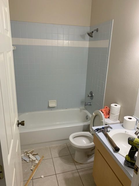 A bathroom featuring a light blue tiled shower area, a white tub, a toilet, and a vanity with tools on the countertop.