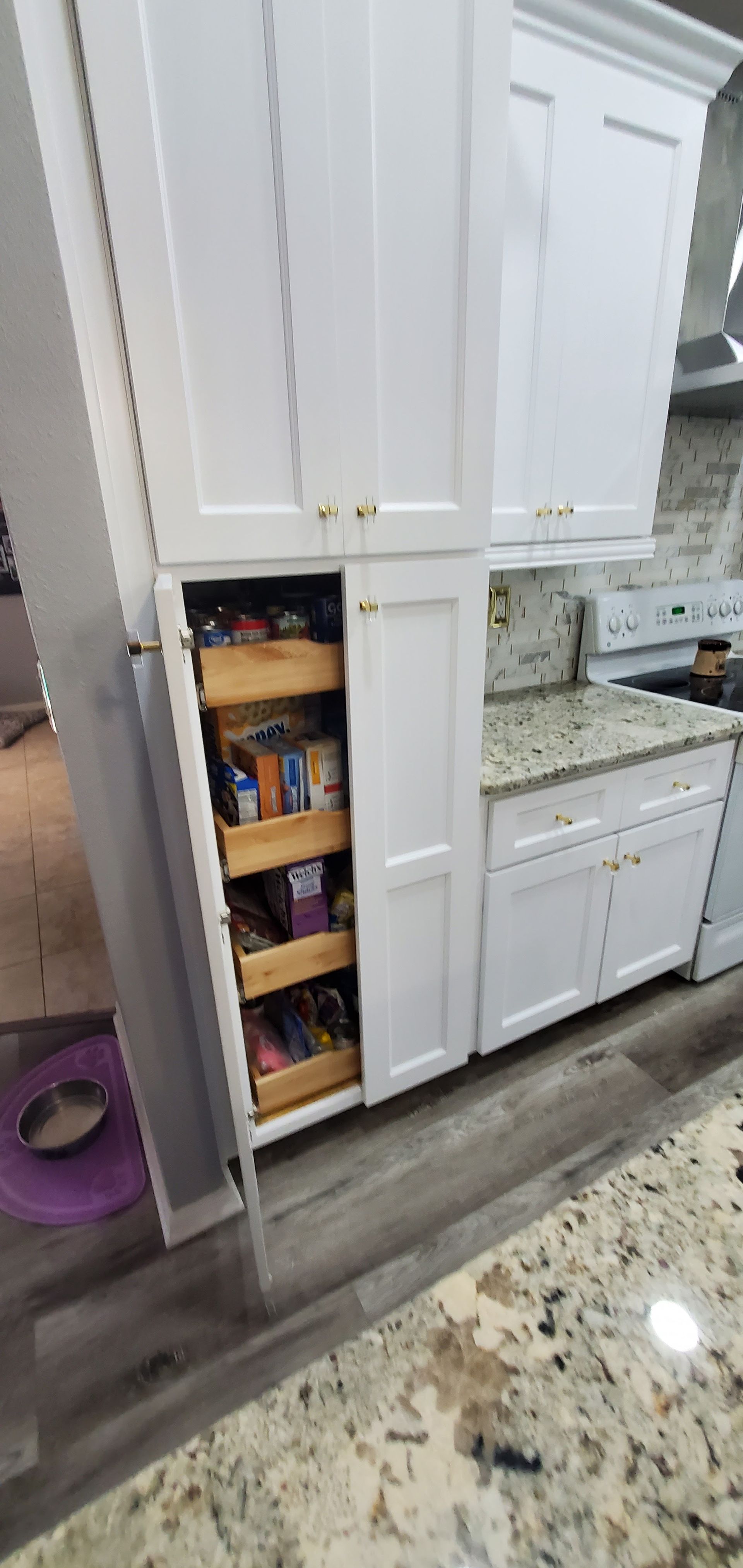 A white kitchen pantry cabinet with open doors revealing several pull-out wooden shelves stocked with various food items.