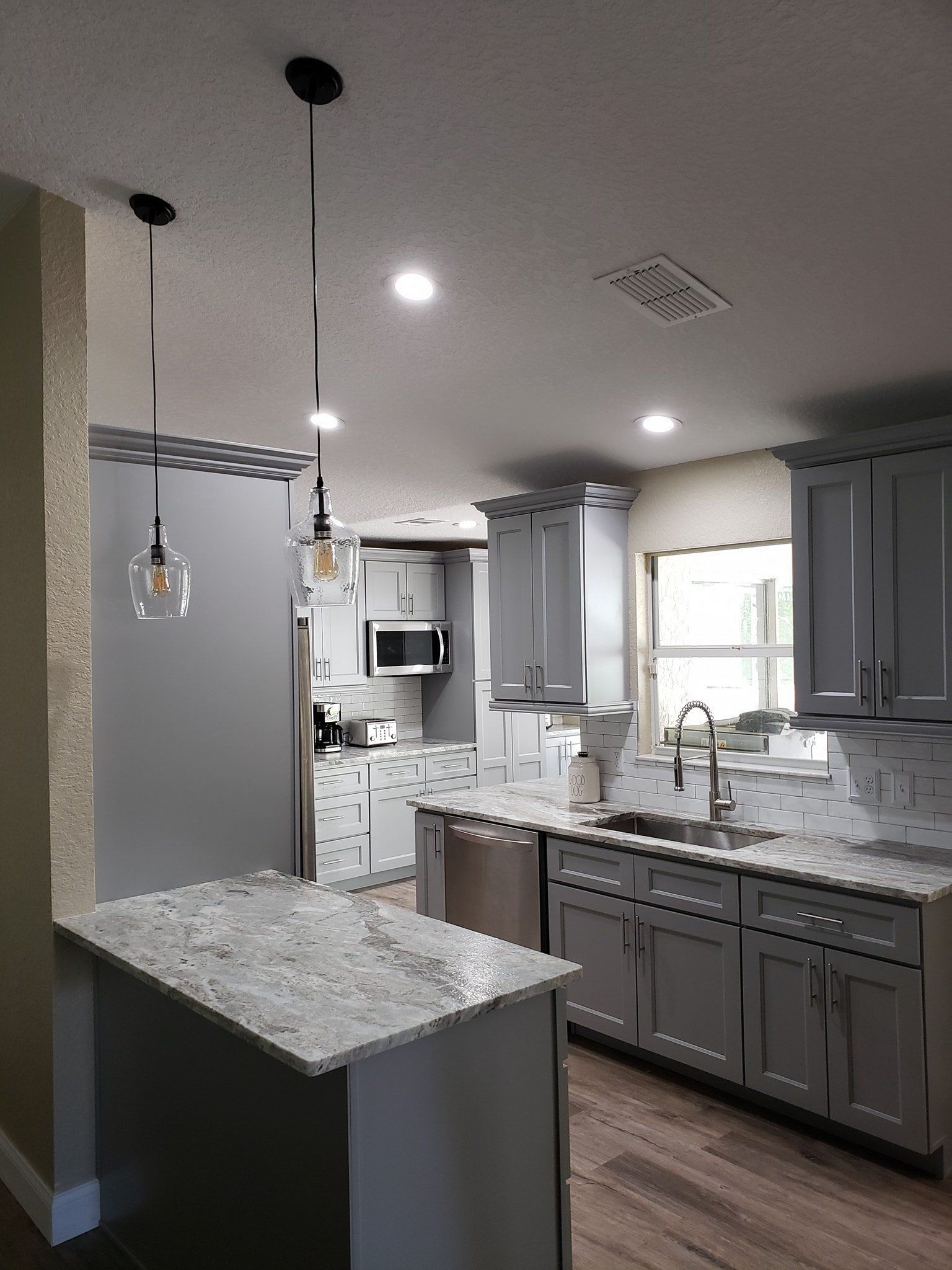 A modern kitchen with grey cabinets, light speckled granite countertops, stainless steel appliances, and pendant lights.