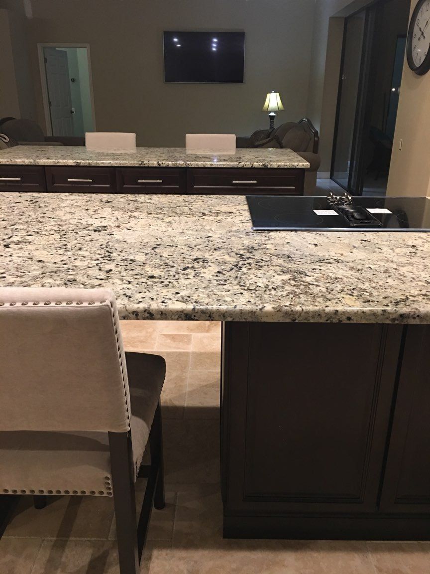 A kitchen island with a speckled granite countertop, dark cabinets, and a beige dining chair in a room with a TV.