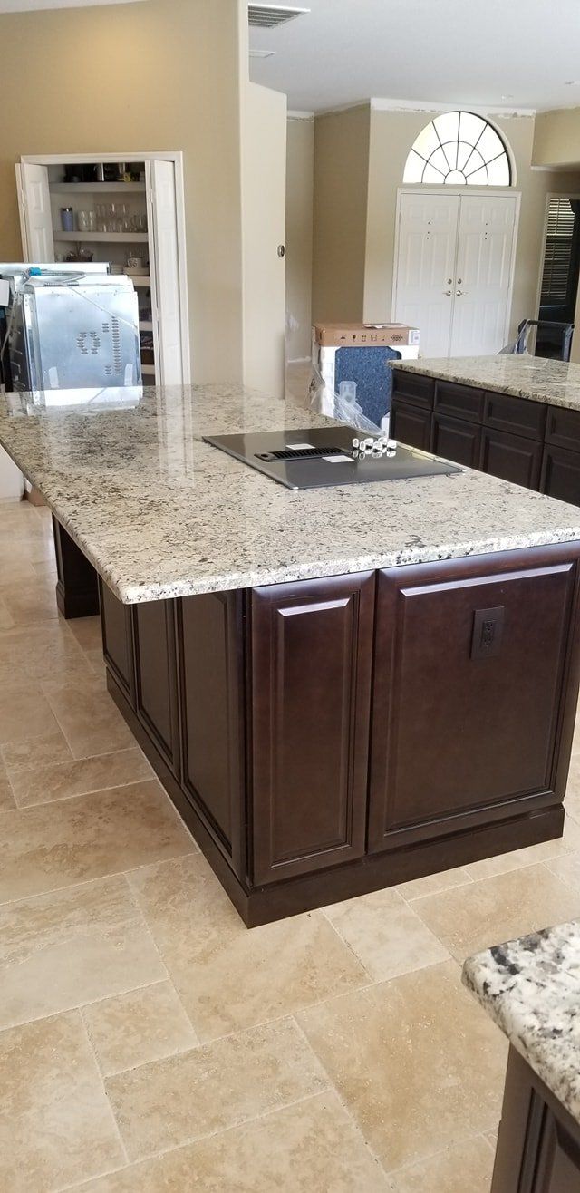 A kitchen island with a granite countertop and dark wood cabinetry on a light tile floor, looking toward a dining room.