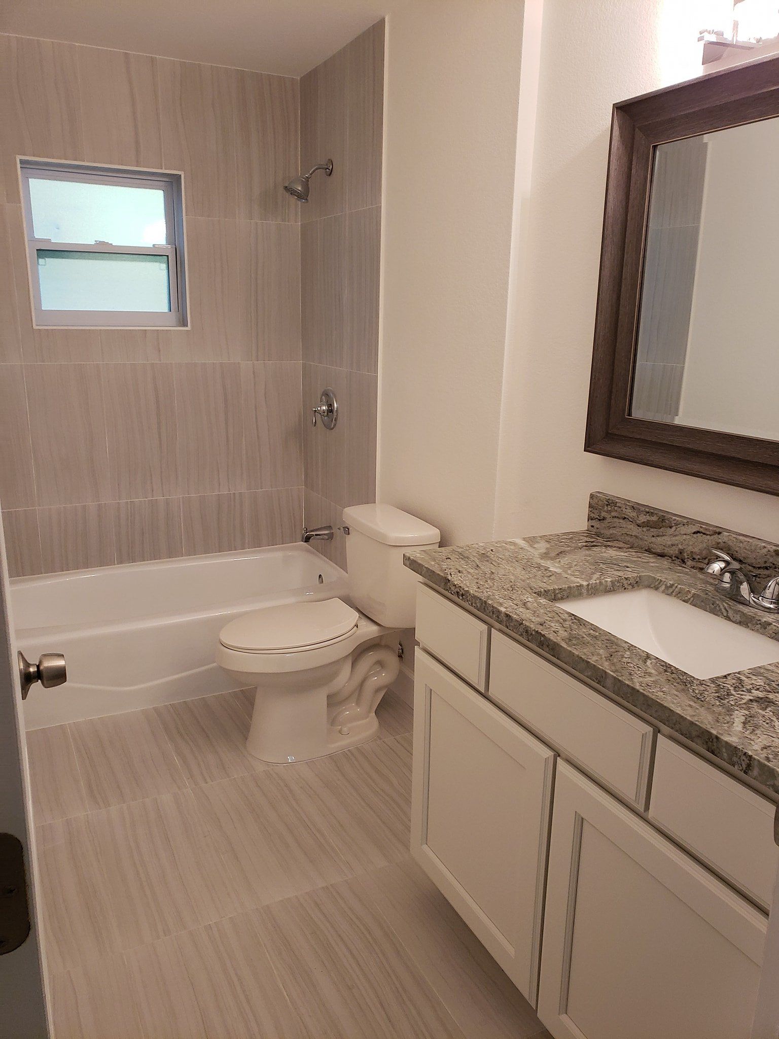 A bathroom with a white toilet, granite-topped vanity, wooden framed mirror, and a tiled shower/tub enclosure.