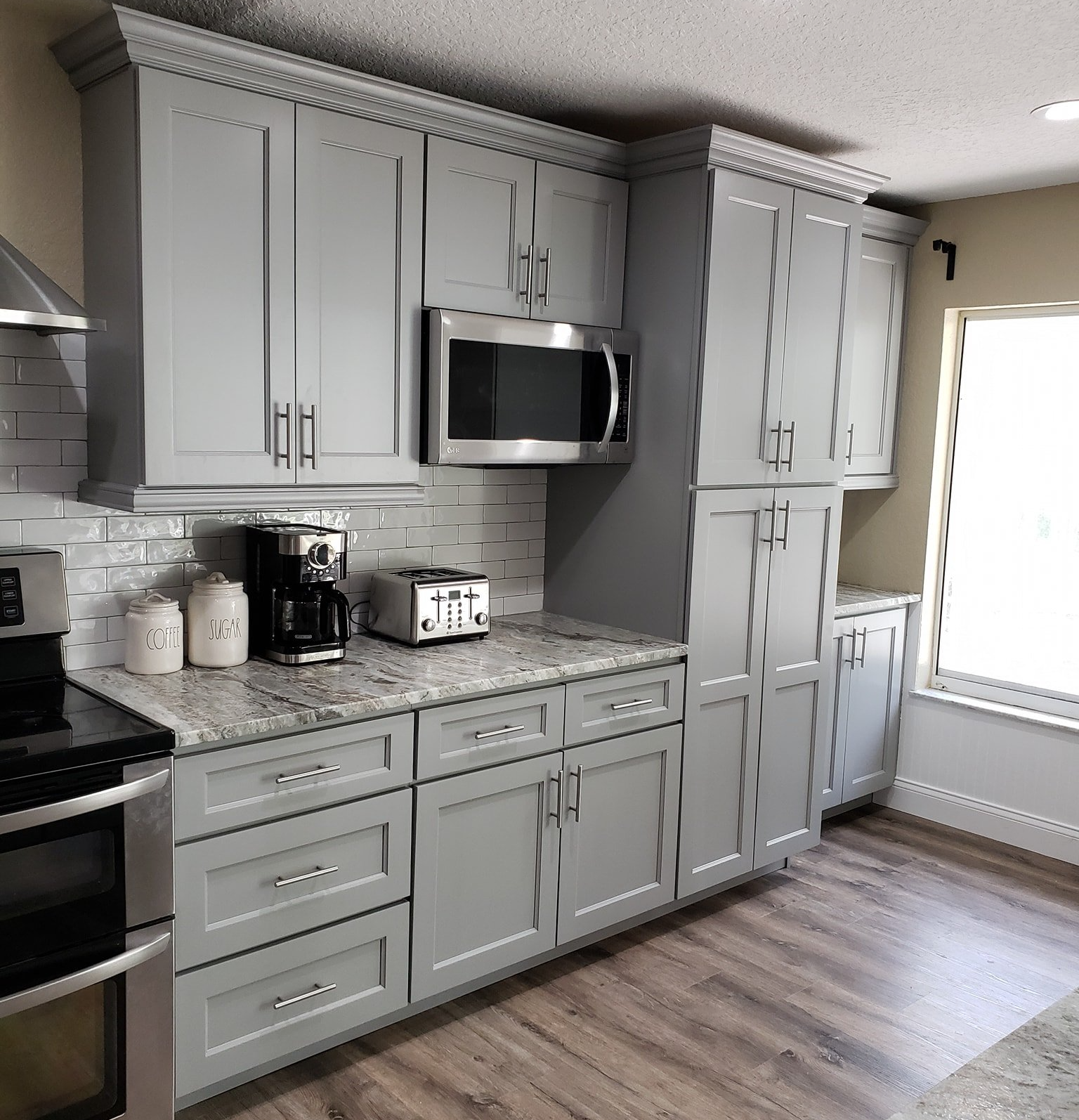 A kitchen interior features grey shaker cabinets, a granite countertop, stainless steel appliances, and wood-look flooring.