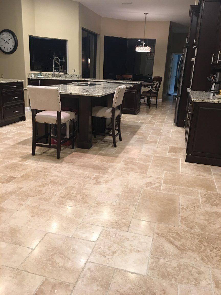 A spacious kitchen with dark wood cabinetry, a granite-topped island with two upholstered chairs, and beige tiled flooring.