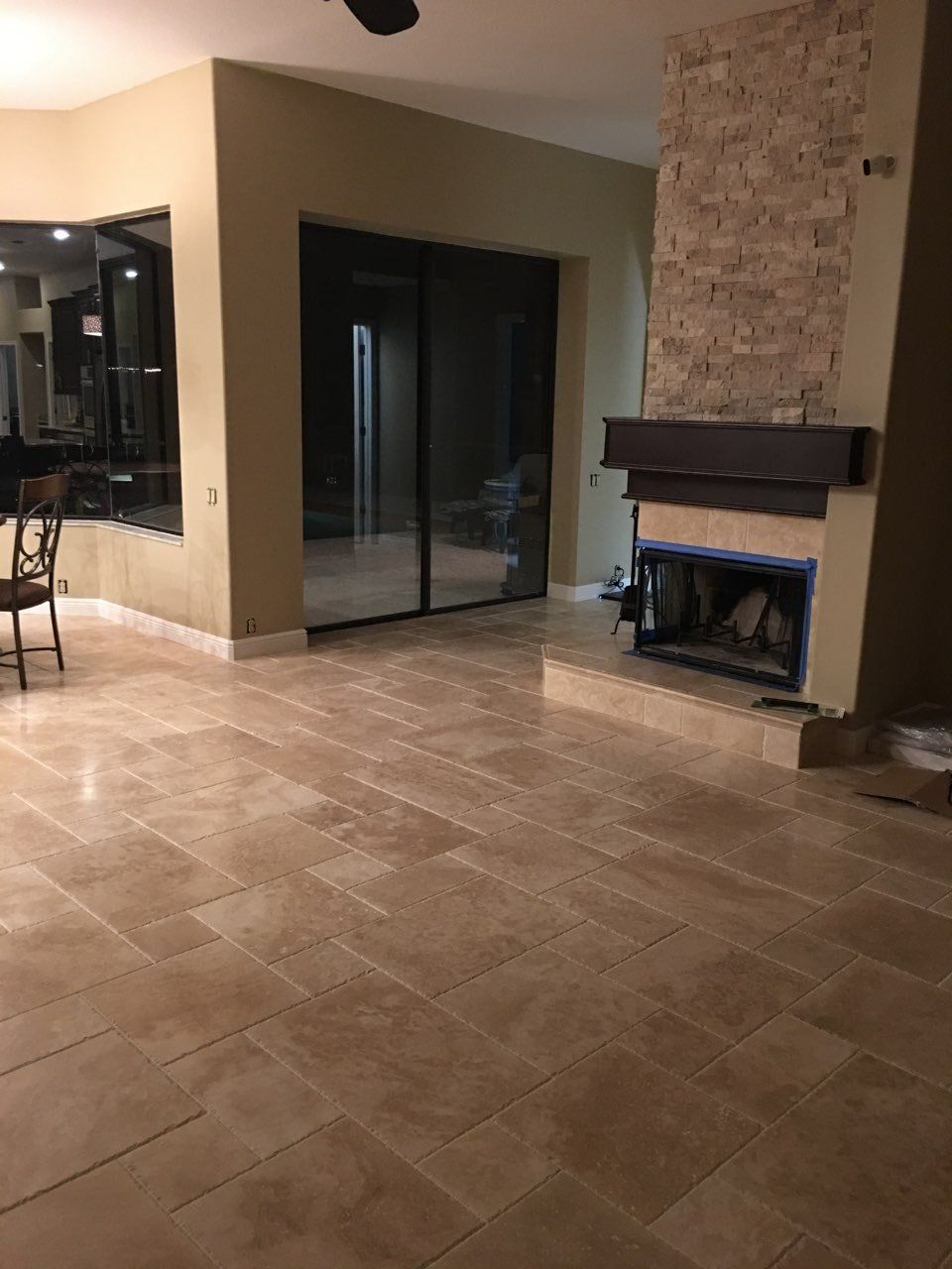 A room with light-colored tile flooring, beige walls, sliding glass doors, and a stone fireplace with a dark mantel.