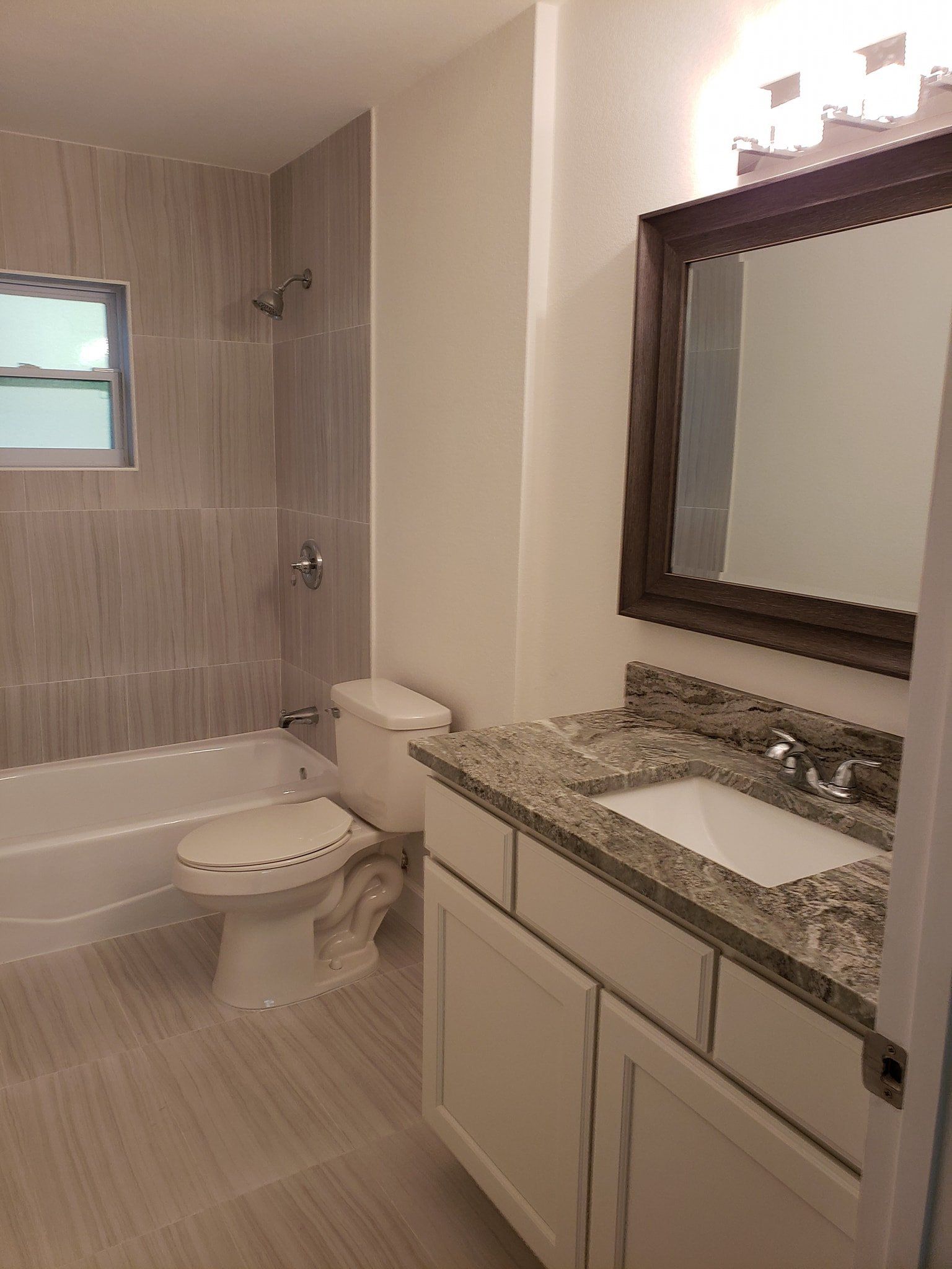 A renovated bathroom with a white vanity, granite countertop, light wood-look tile, a toilet, and a bathtub with shower.