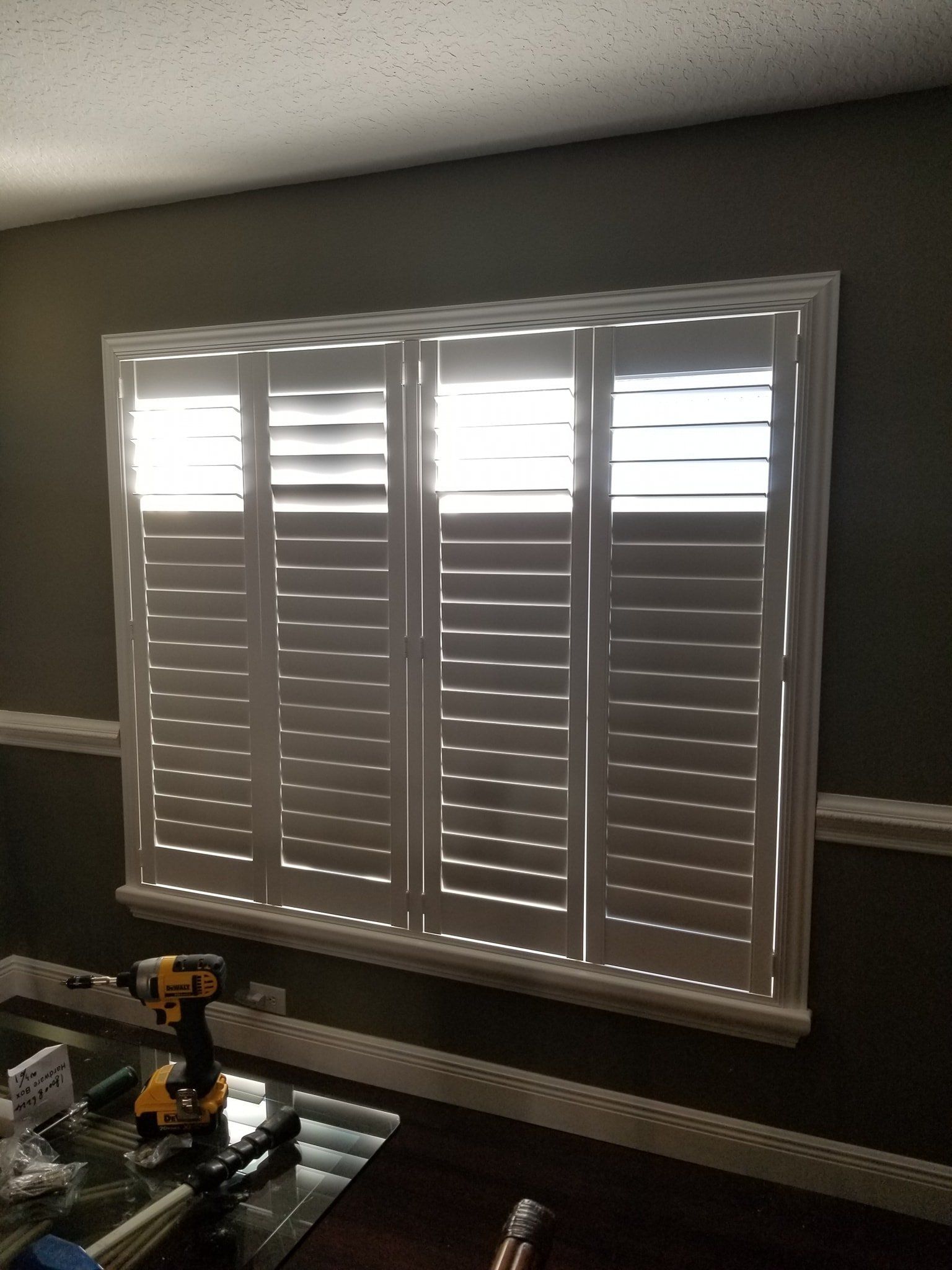 White plantation-style shutters installed on a window in a room with dark olive walls and white trim.