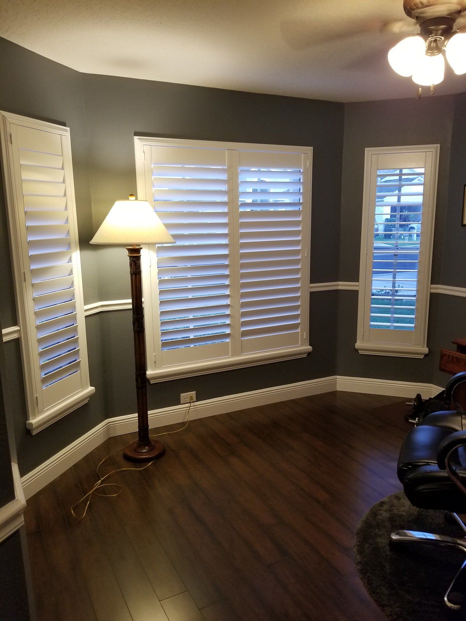 A room with dark gray walls, white plantation shutters on three windows, dark wood floors, and a floor lamp.
