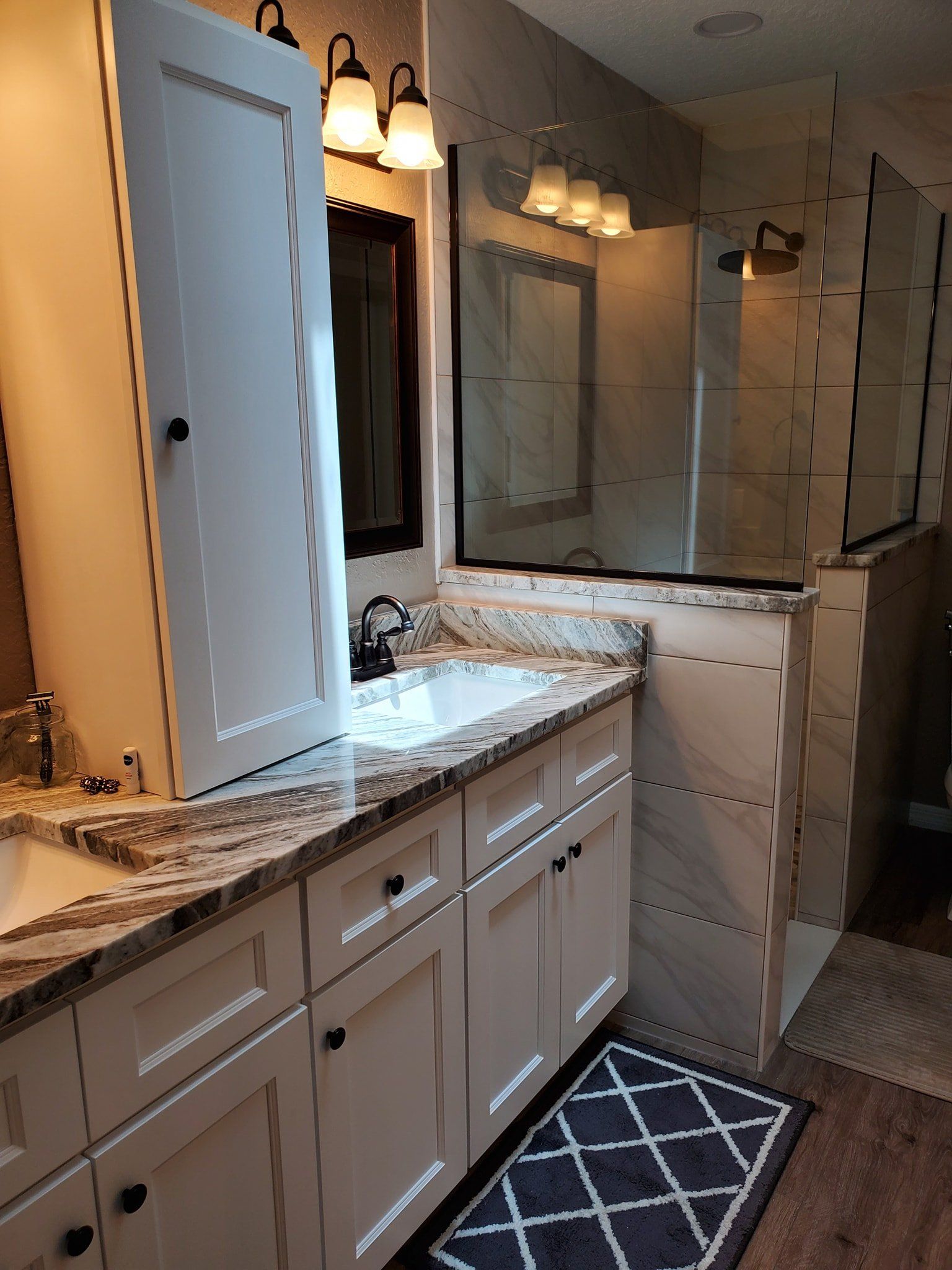 A bright bathroom vanity with white cabinets, granite countertops, and a glass-enclosed shower area.