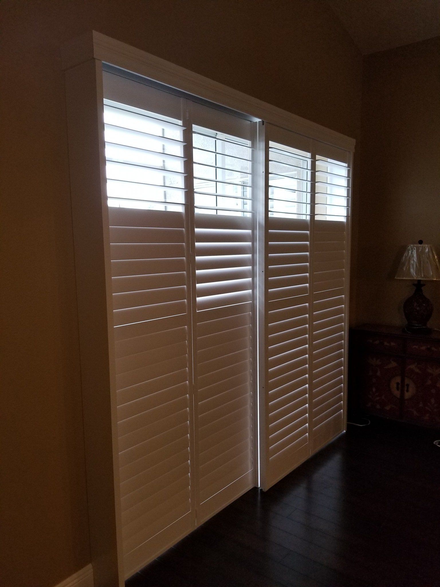 White louvered plantation shutters cover a large window in a room with dark wood flooring and a table lamp.
