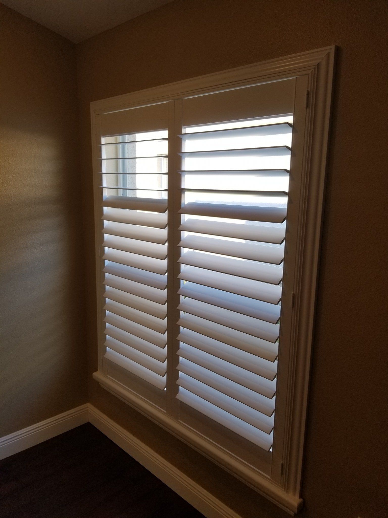 White interior plantation shutters installed on a window in a room with brown walls and a dark floor.