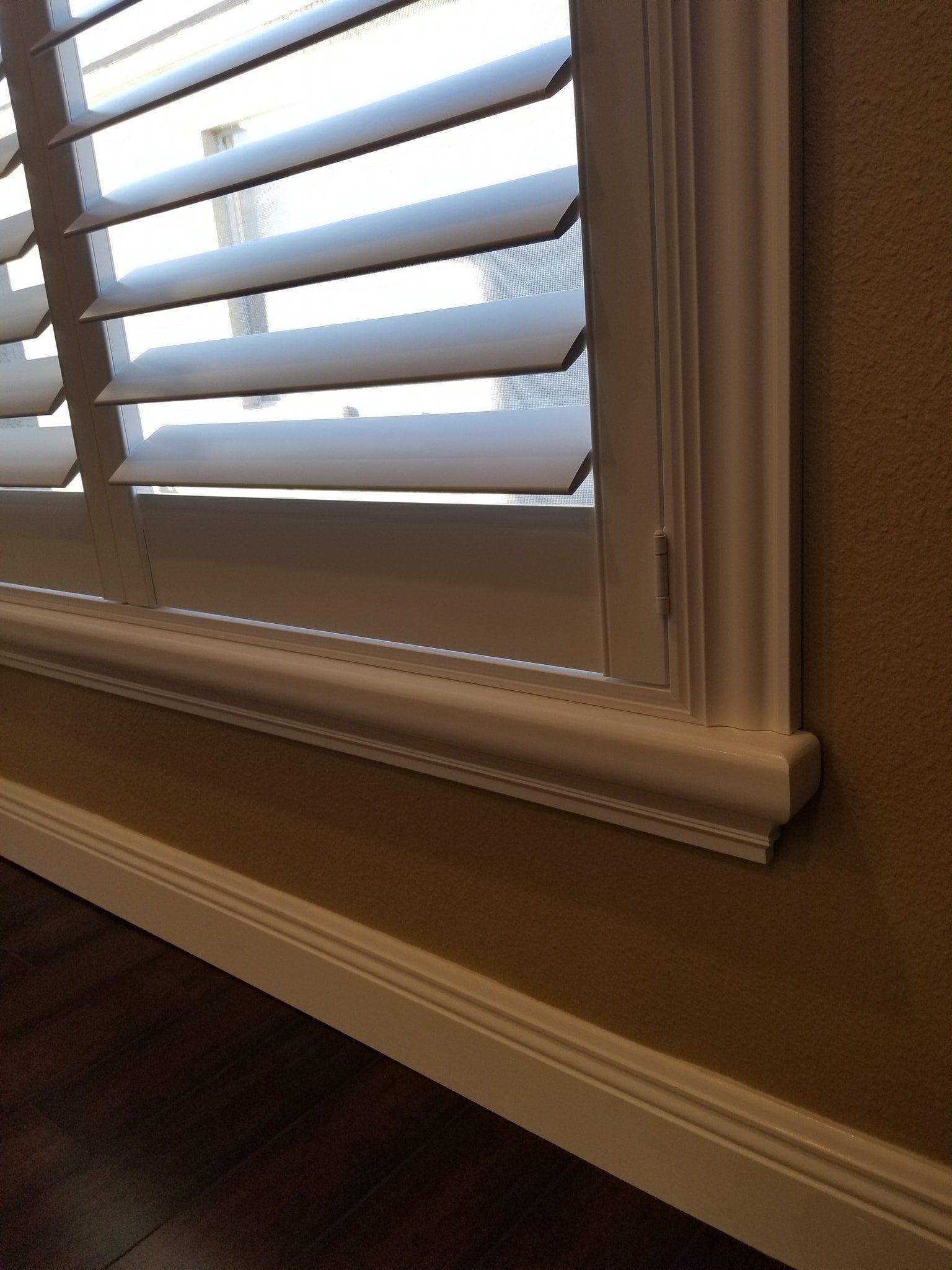 White plantation shutters installed on a window with a decorative white wooden sill above a baseboard against a tan wall.