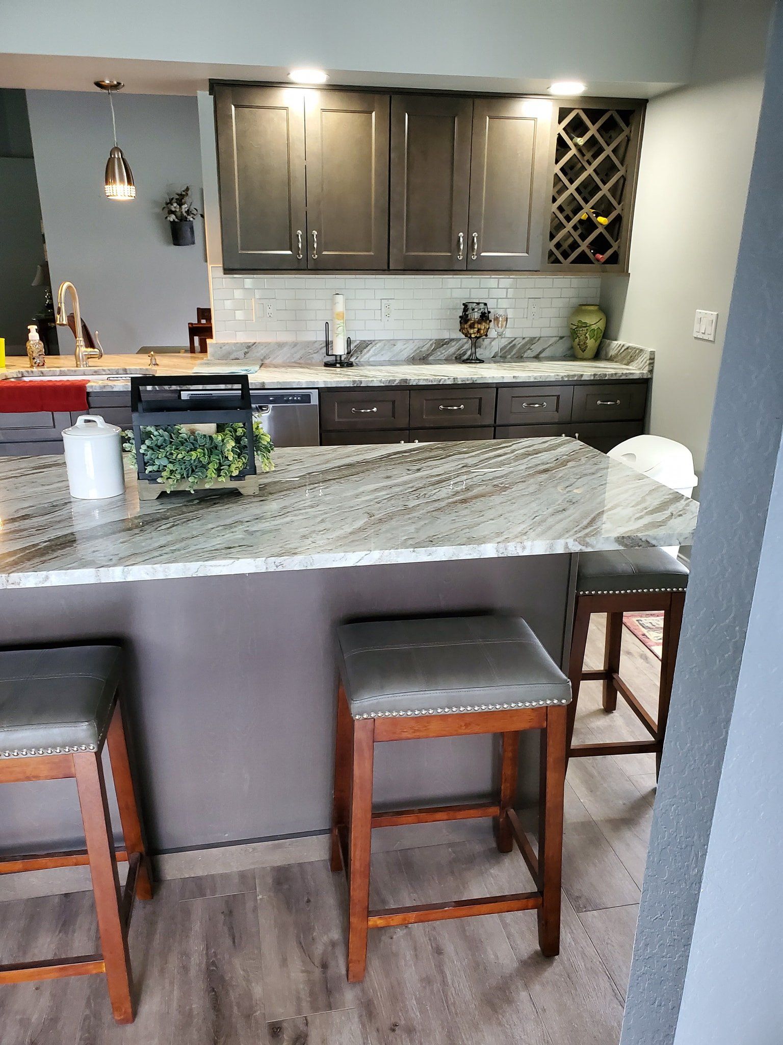 A kitchen island with two stools in the foreground, facing dark cabinets, a white tile backsplash, and granite counters.