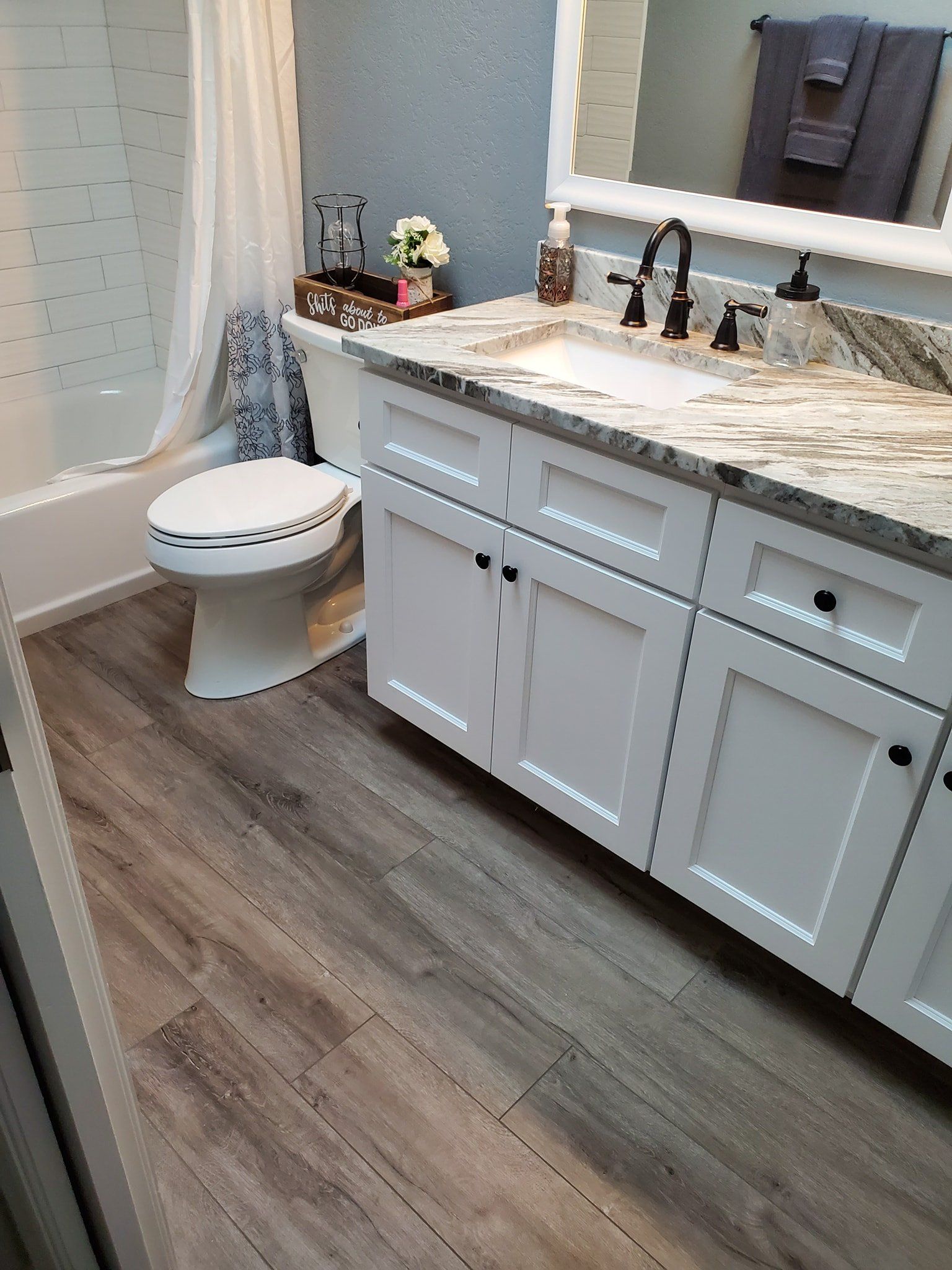 A modern bathroom featuring a white vanity with granite countertops, light gray cabinets, and wood-look laminate flooring.