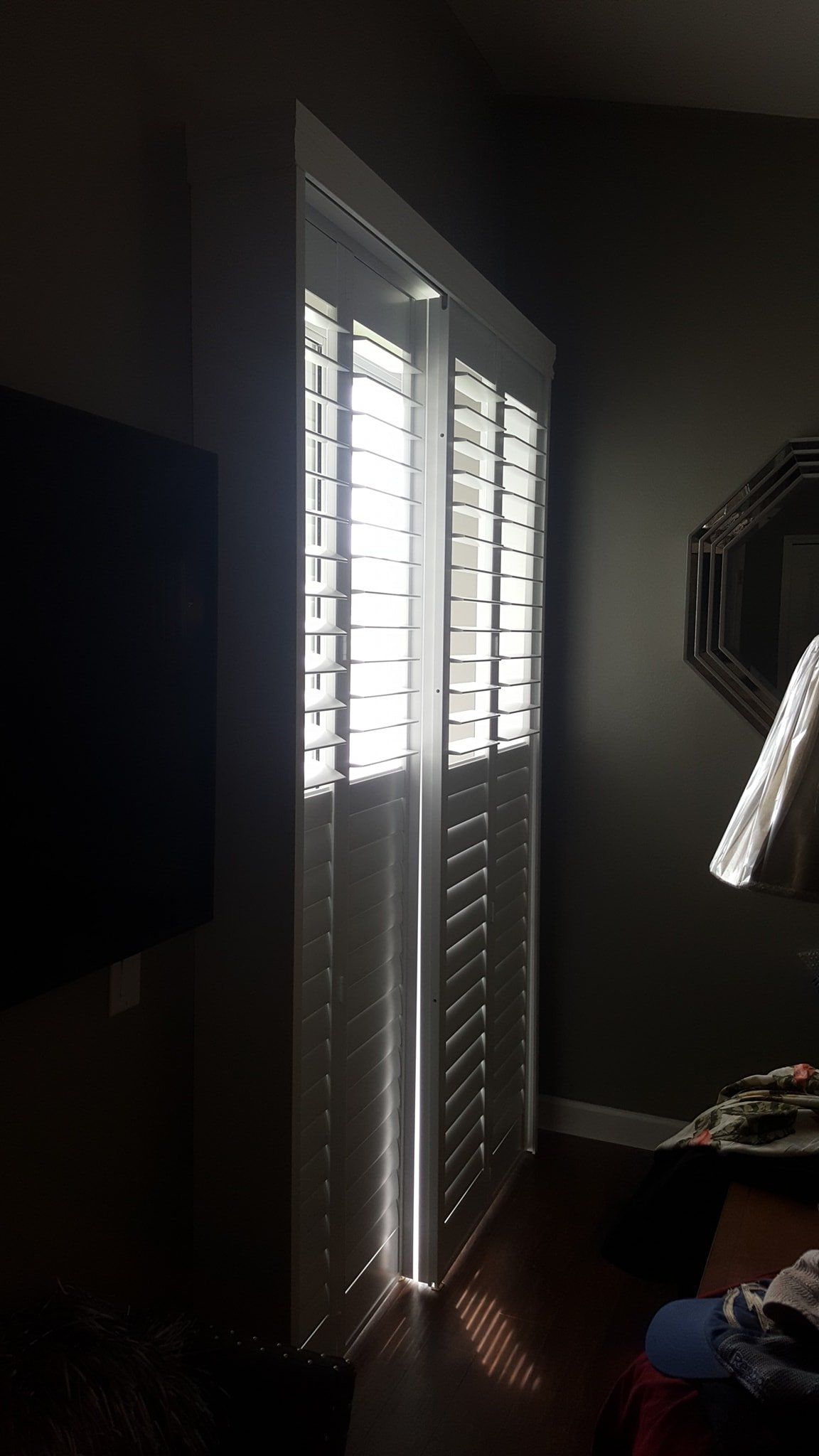 White indoor plantation shutters on a tall window, with bright daylight streaming through the slats into a dark room.