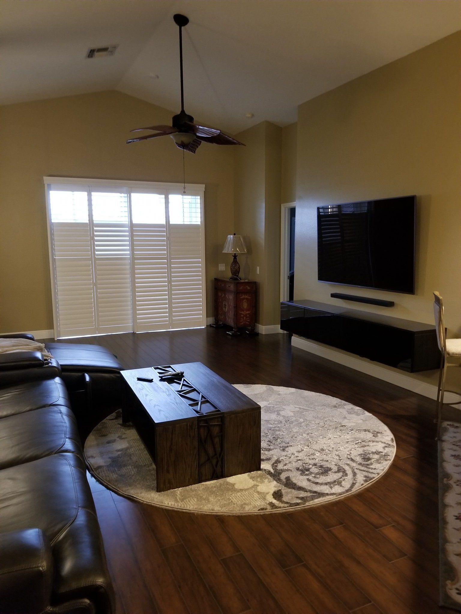 A living room with a large TV mounted on a beige wall, a dark wood coffee table on a circular rug, and glass patio doors.
