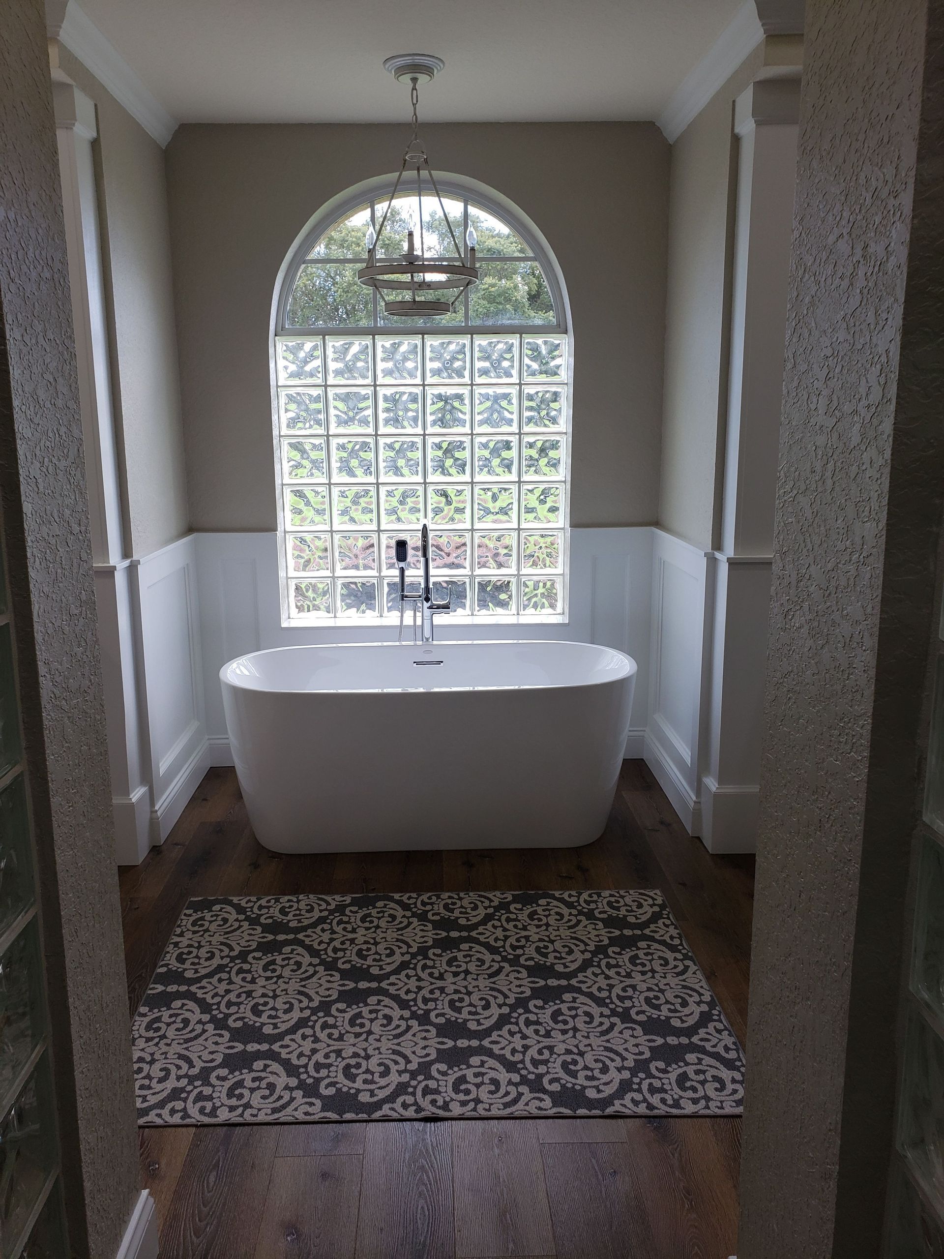 A white soaking tub centered beneath an arched glass-block window in a bathroom with hardwood floors and wainscoting.