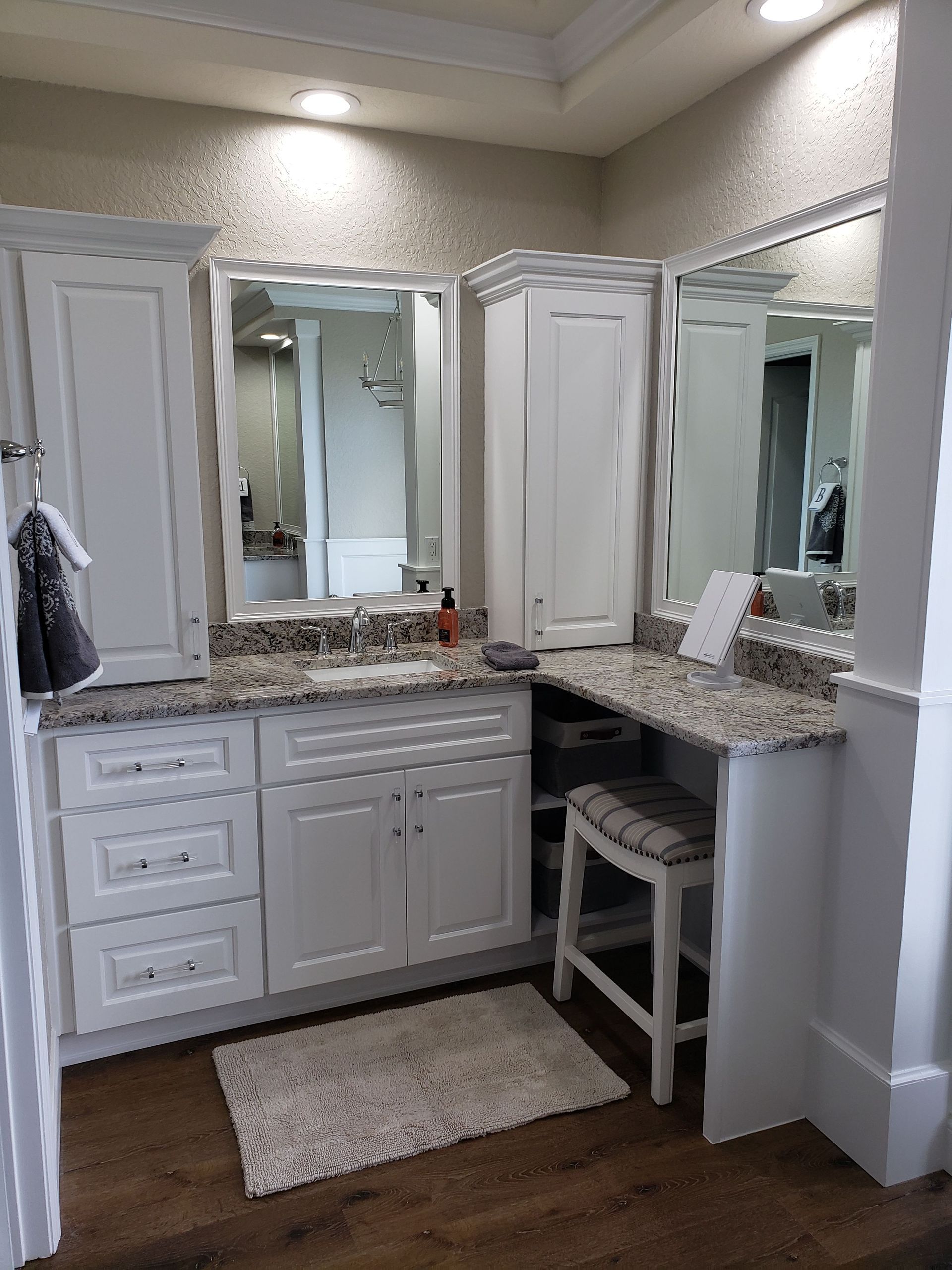 A bathroom vanity with white cabinets, granite countertops, two mirrors, a matching stool, and a beige rug on wood floors.