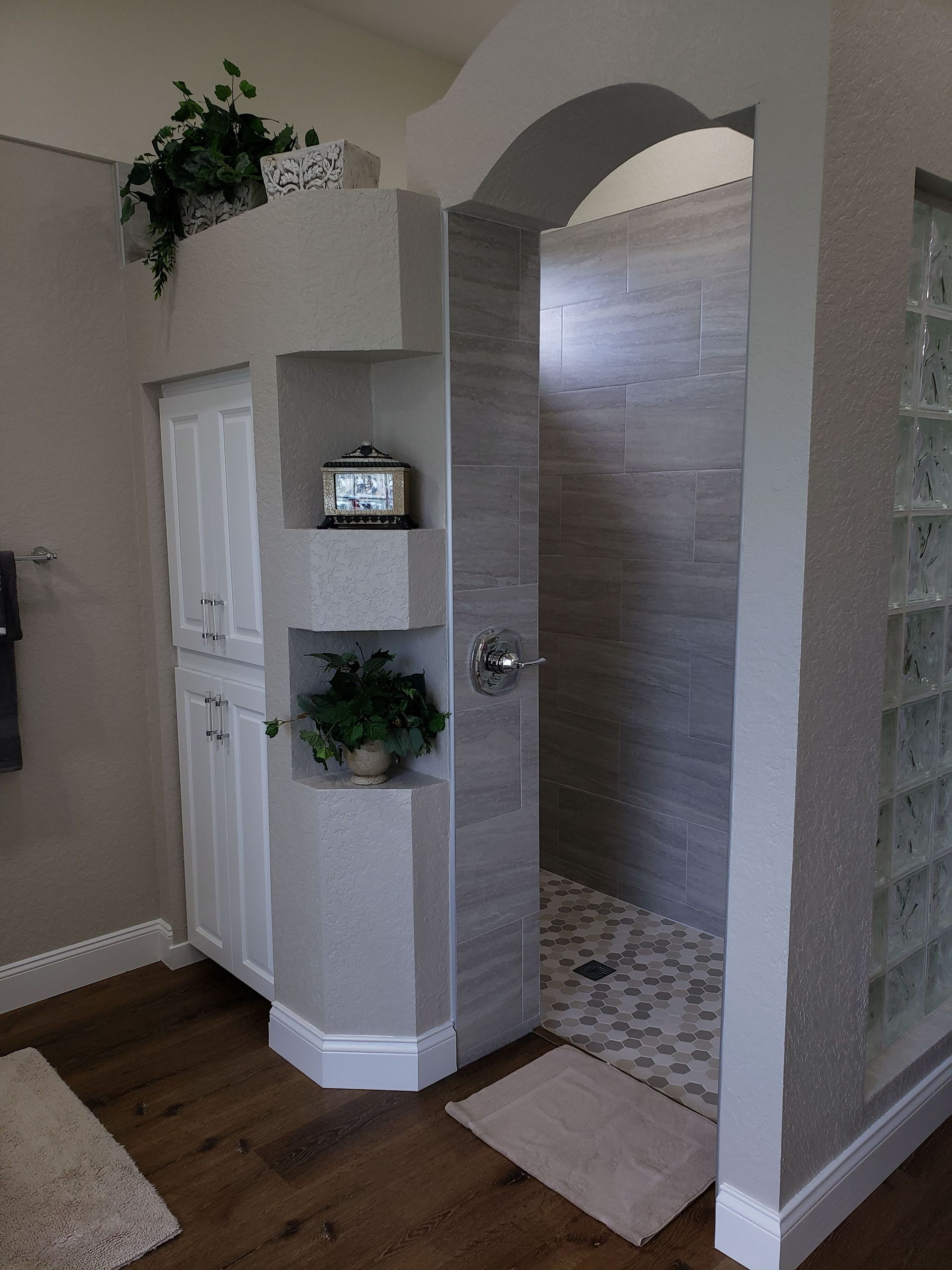 A walk-in shower with a beige arched entry, grey stone walls, pebbled floor, and an adjacent white cabinet with shelves.