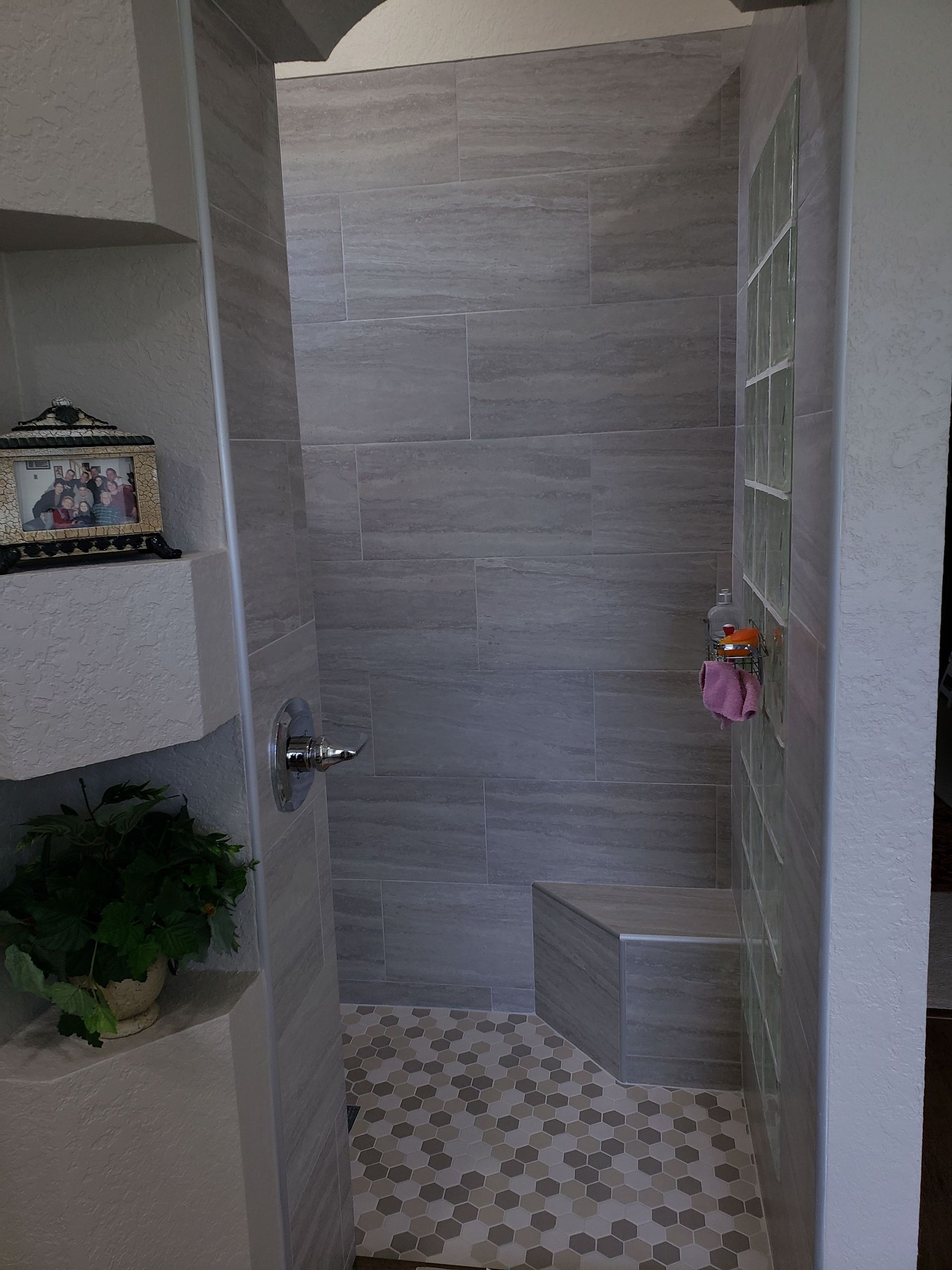 A shower stall with gray tiled walls, a pebble-style floor, a corner bench, and glass block accent tiles.