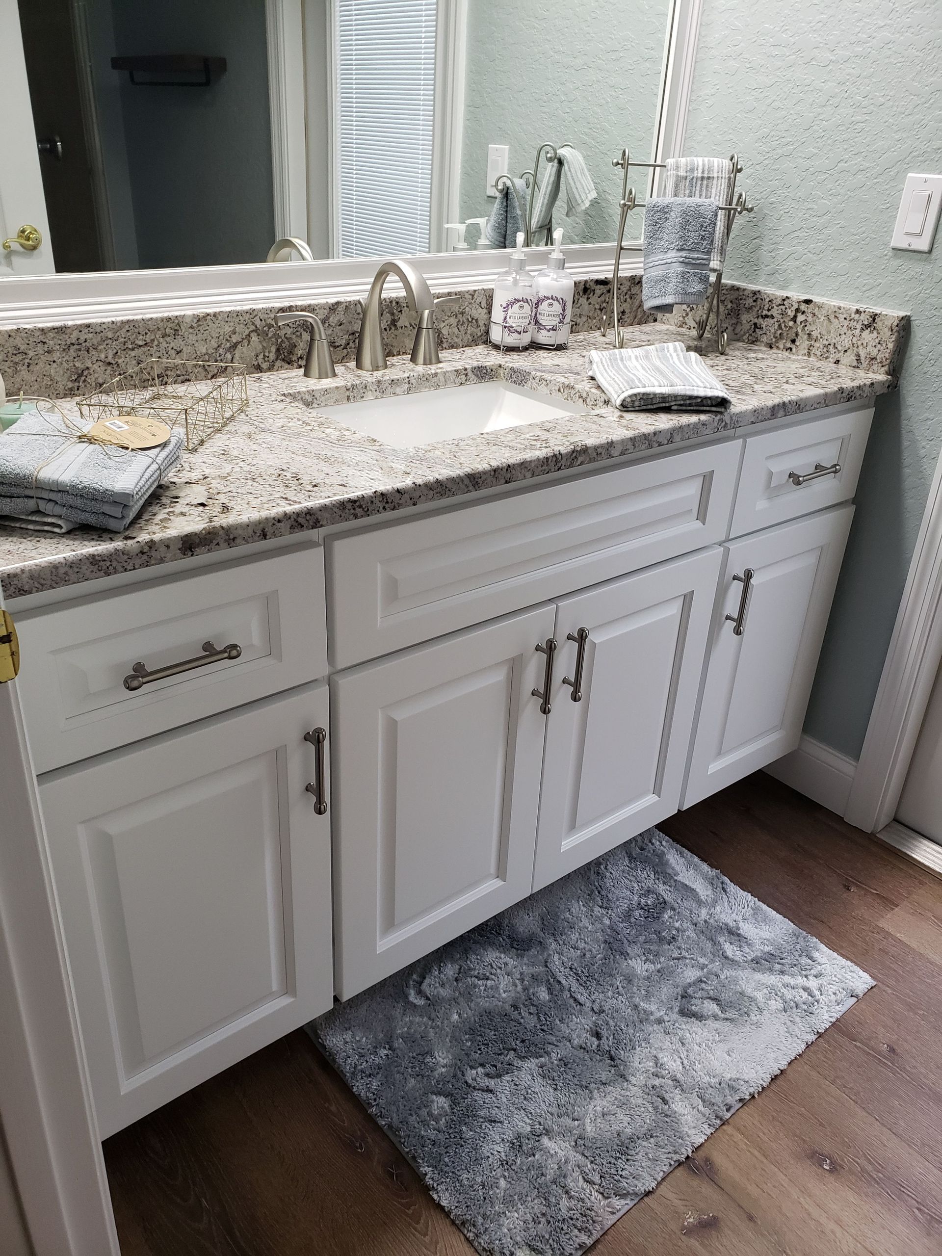 White vanity cabinet with granite countertop, sink, faucet, and mirror in a bathroom with patterned wallpaper.