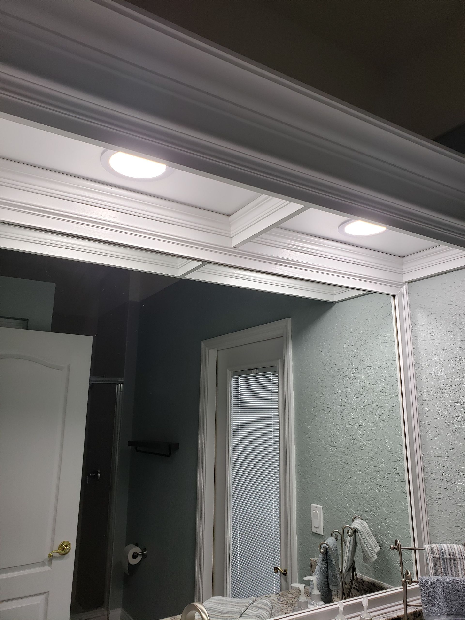 A bathroom mirror featuring intricate white molding, framed by decorative trim with two overhead recessed lights.