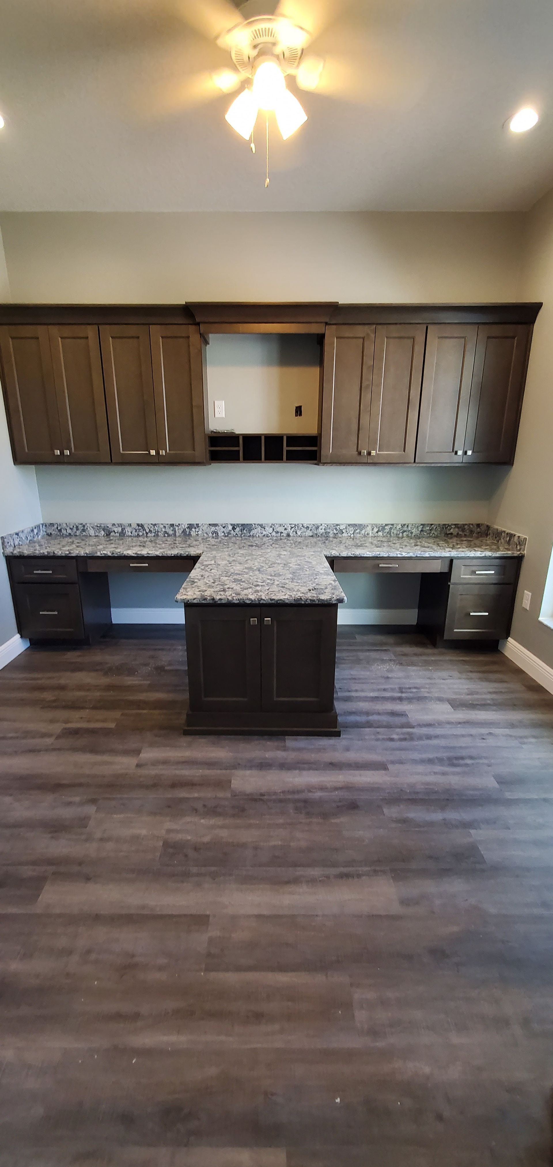 A home office with dark wood cabinetry, granite countertops, a center desk island, and gray wood-look plank flooring.