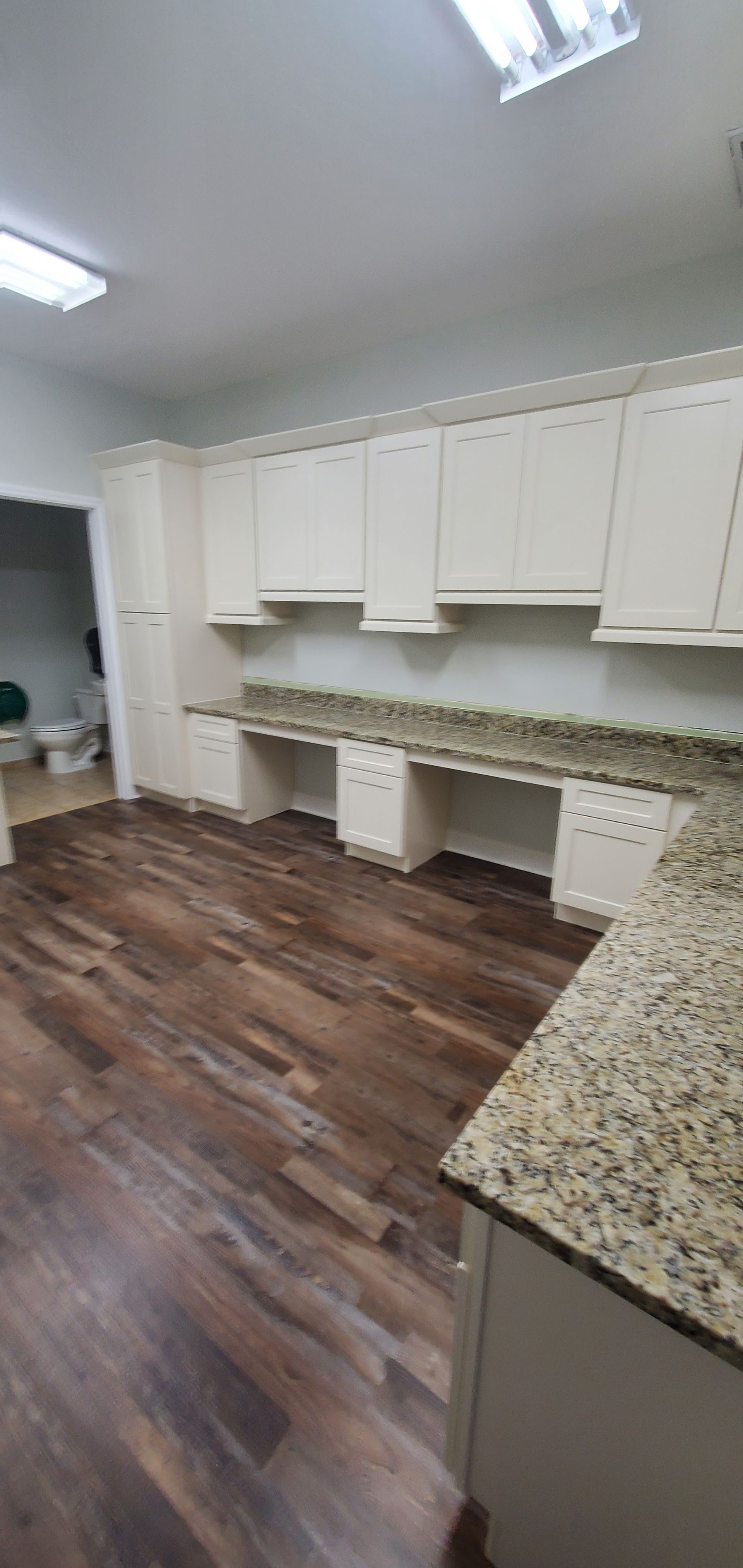 A kitchen with cream cabinets, granite countertops, and dark wood-look flooring, showing an open desk-like workstation.