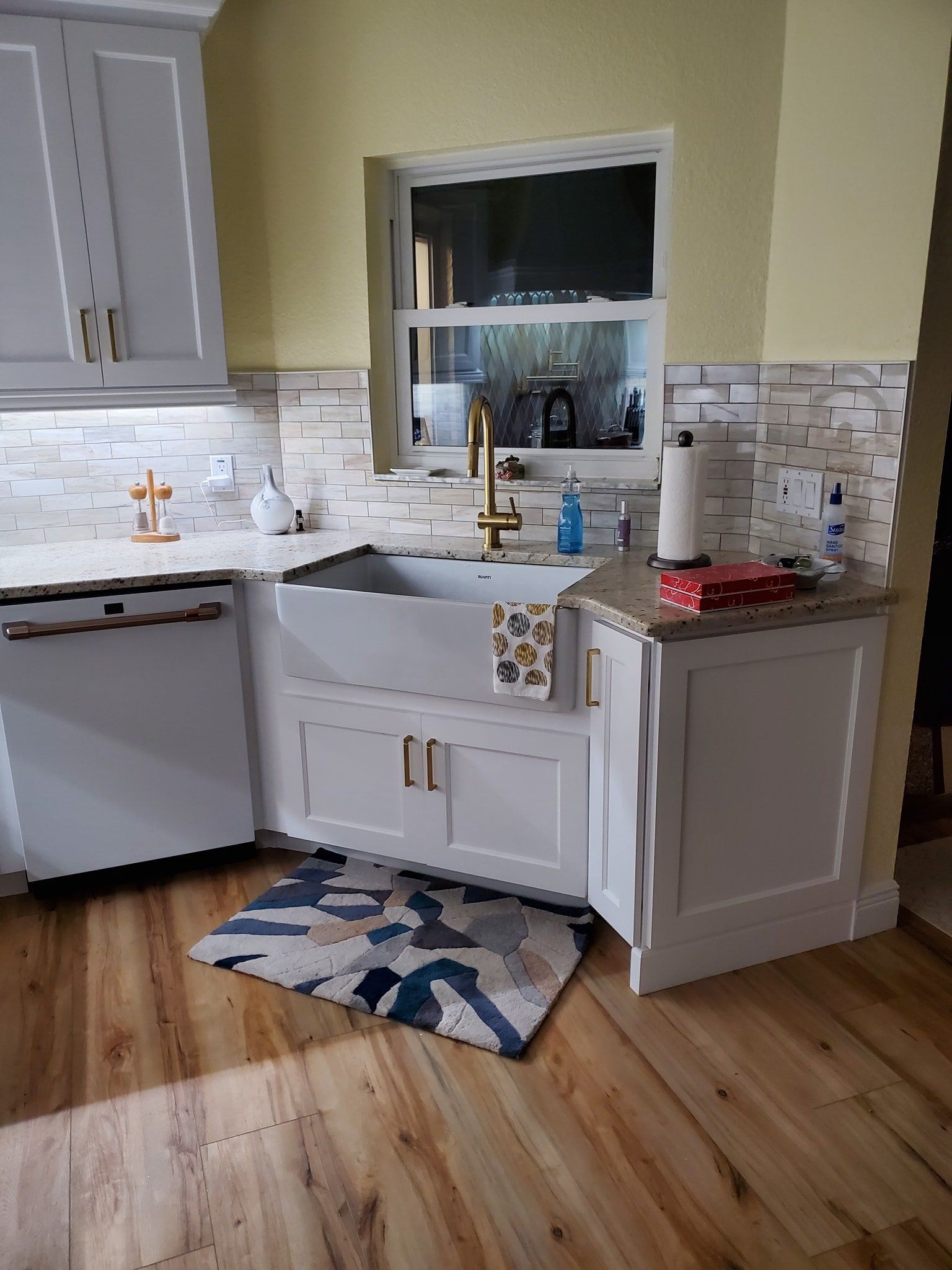 A modern kitchen with white cabinets, a farmhouse sink, brass faucet, light wood flooring, and patterned kitchen rug.