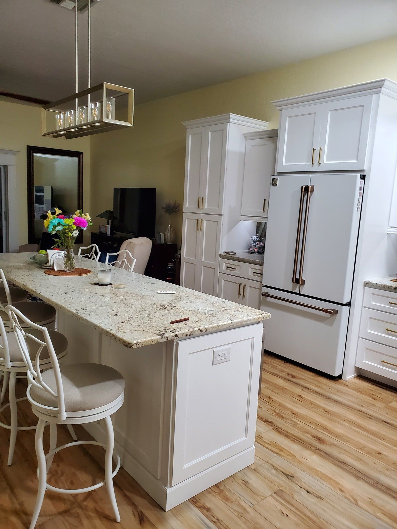 A white kitchen island with granite countertops, bar stools, and a matching refrigerator in a light-walled room.