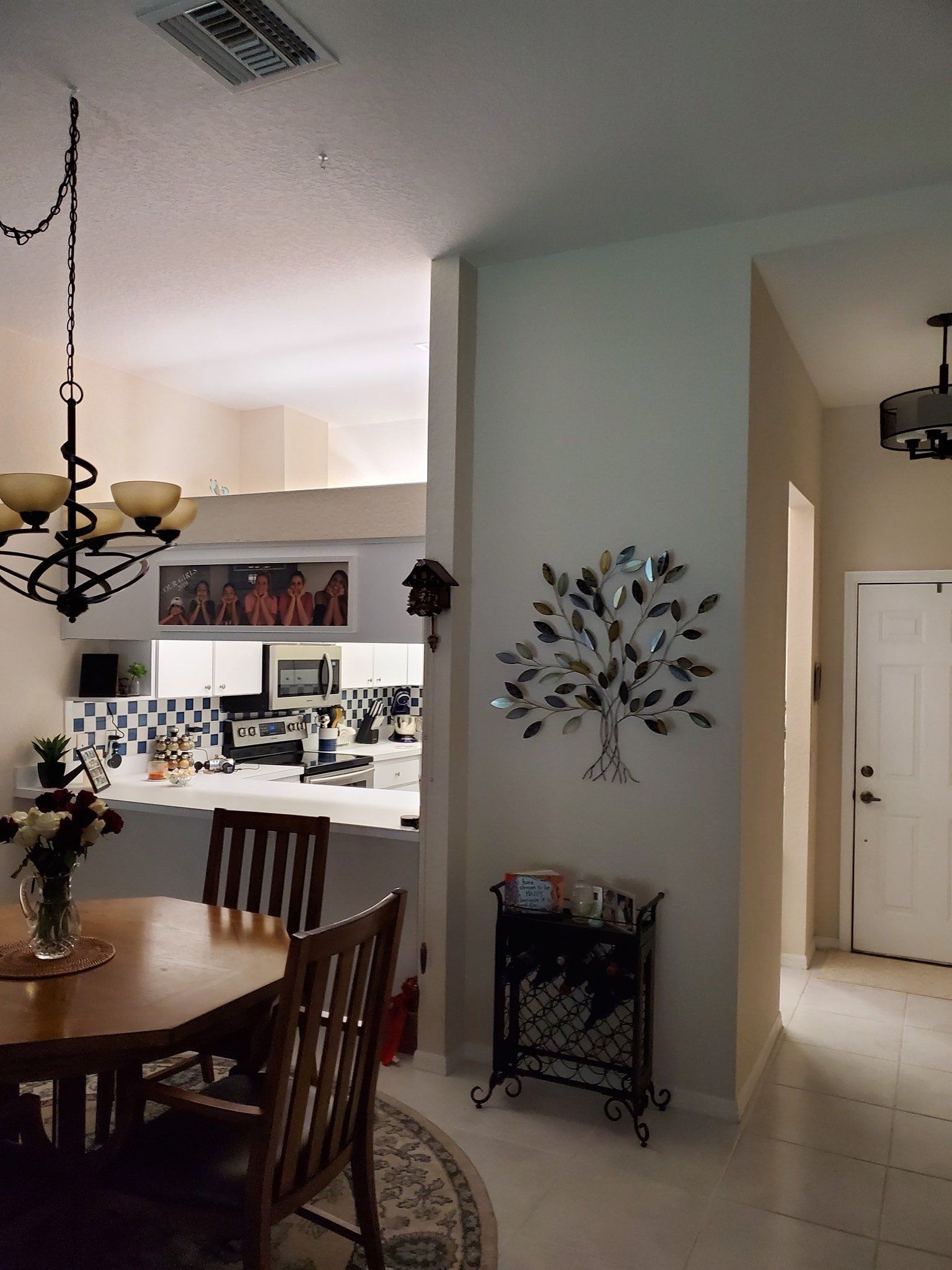 A dining area with a round wooden table, a kitchen visible in the background, a metal tree wall sculpture, and a doorway.