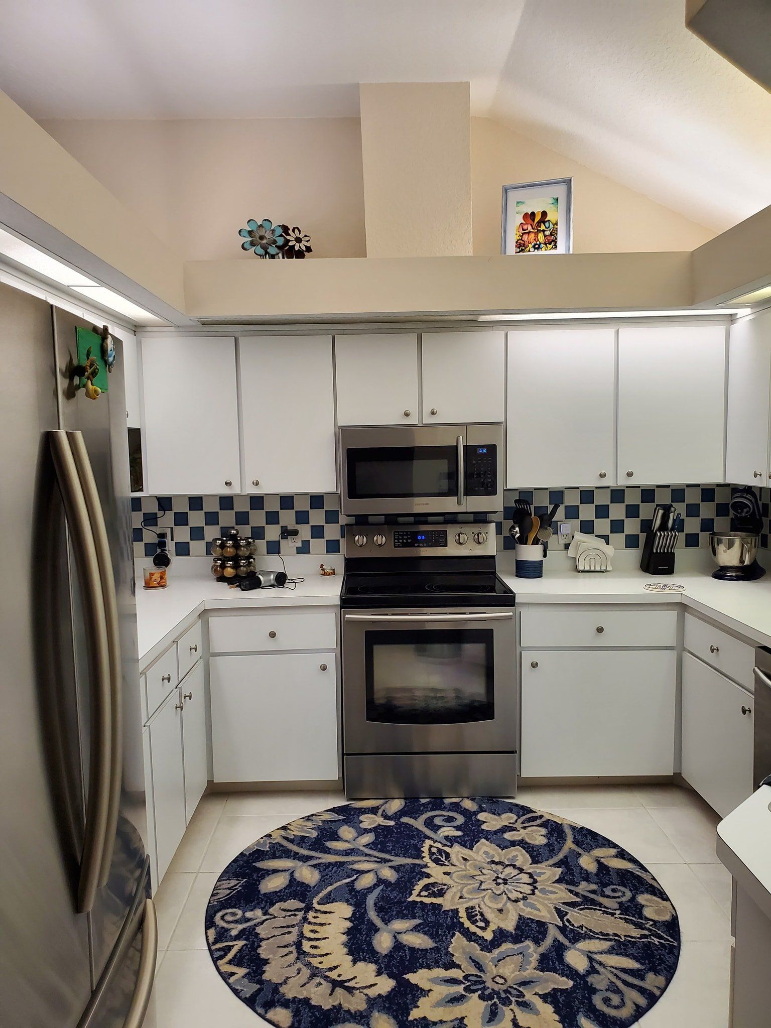 A kitchen with white cabinets, stainless steel appliances, a checkered backsplash, and a blue floral round area rug.