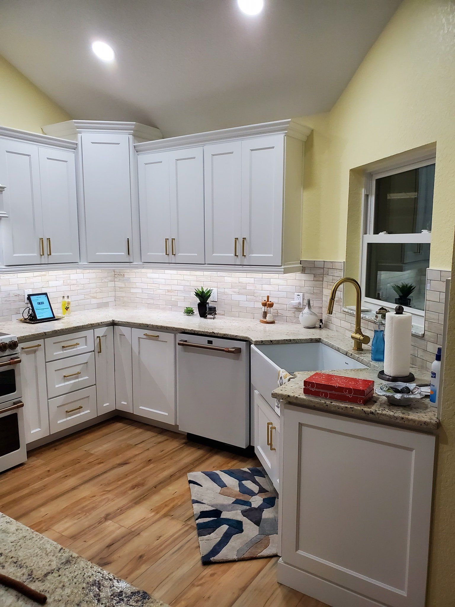 A brightly lit kitchen with white cabinets, granite countertops, light wood floors, and a farmhouse sink.