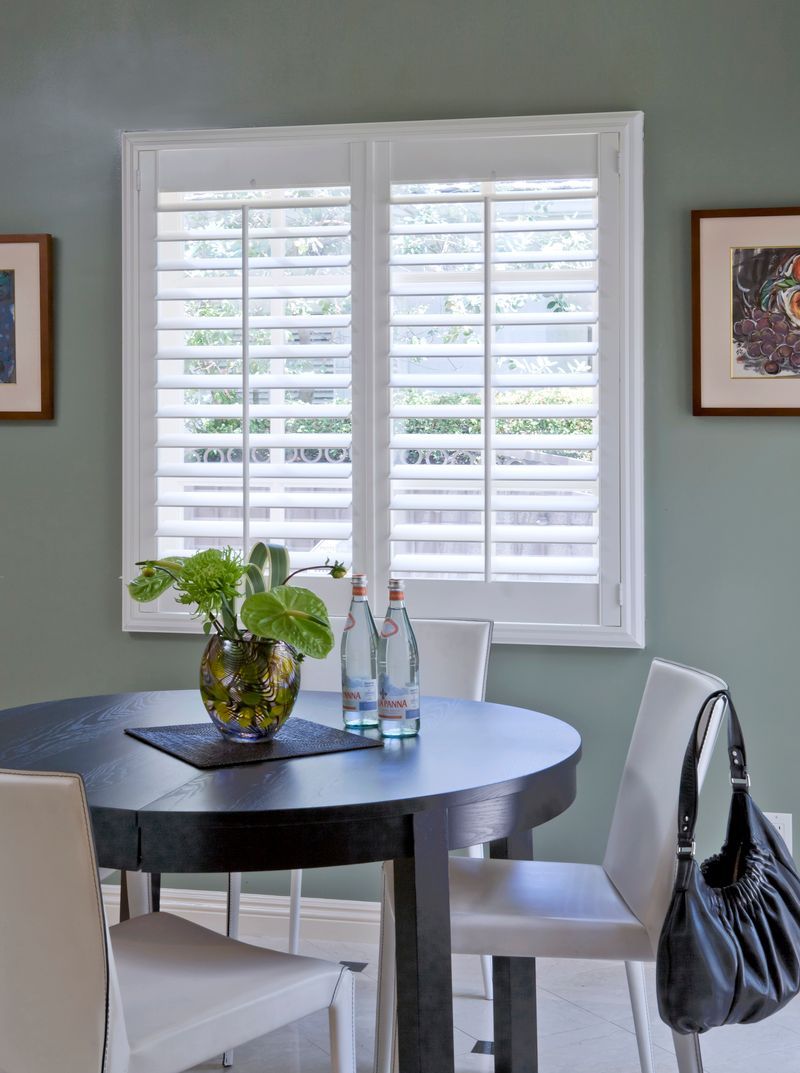 A round, dark-finished dining table with a green plant, two water bottles, and white chairs in a room with shuttered windows.