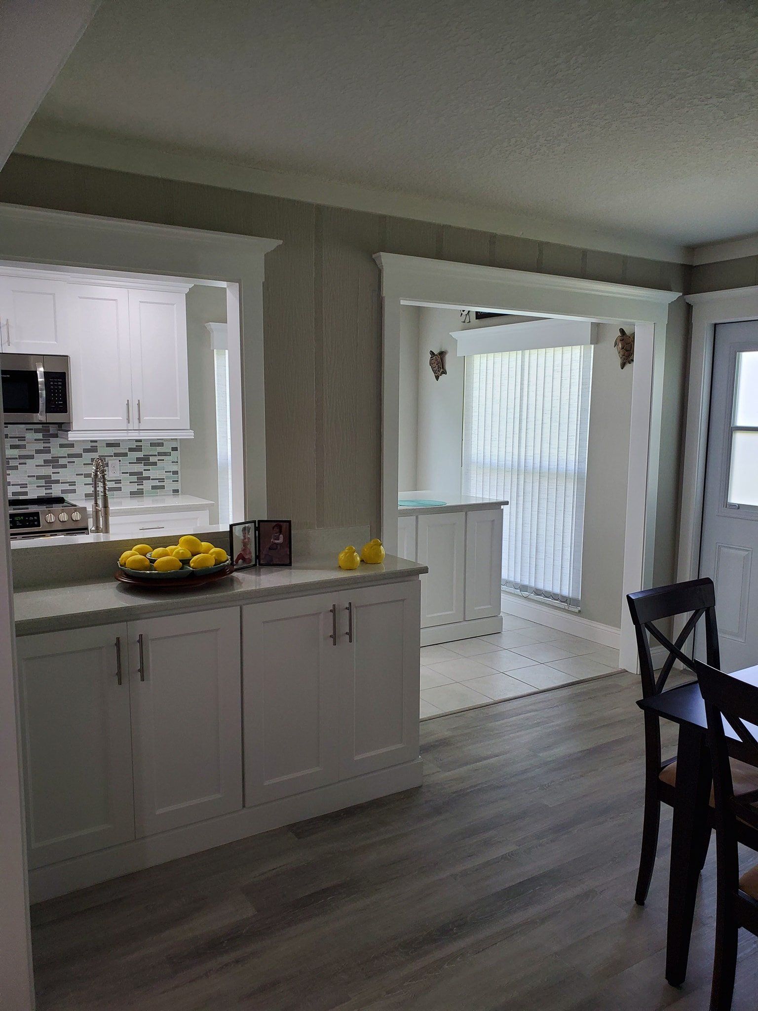 A bright kitchen features white cabinets, a light gray countertop with a bowl of lemons, and wide doorways to a dining area.